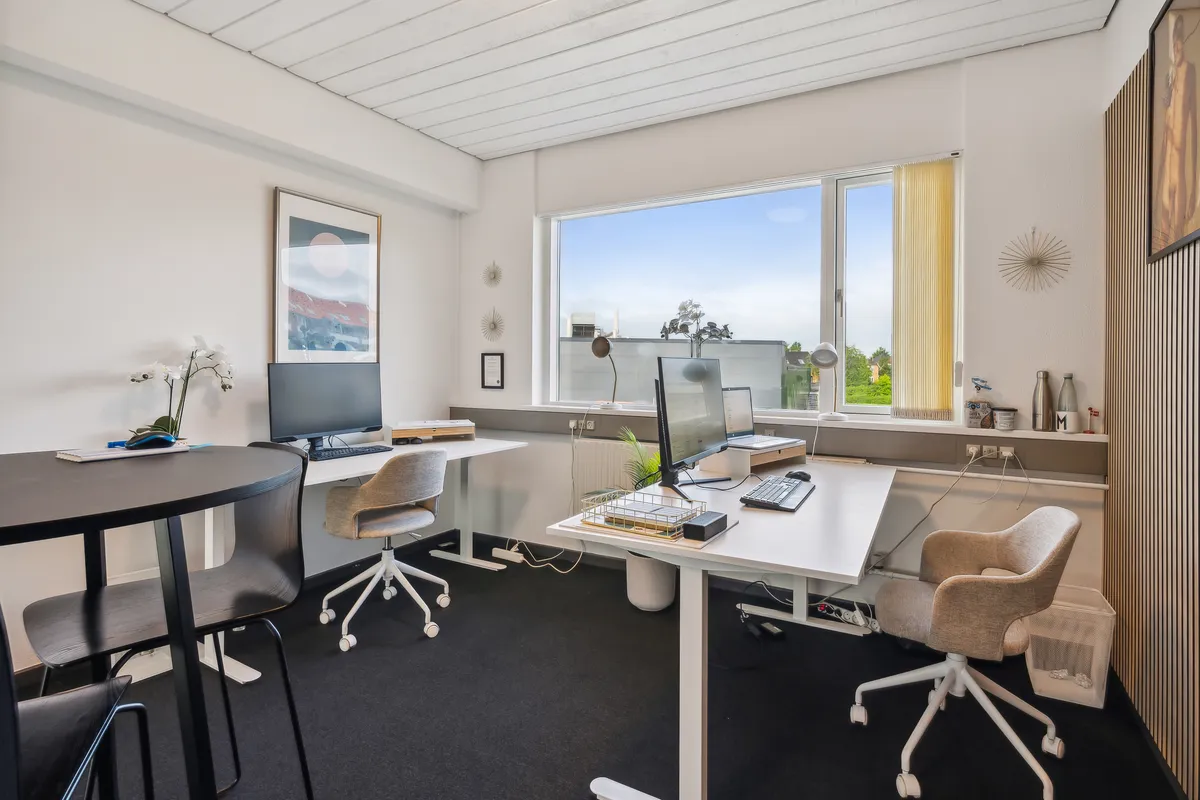 Kontor med moderne møbler, høje vinduer, hvide vægge, to skriveborde og en separat arbejdszone med plante og to skærme