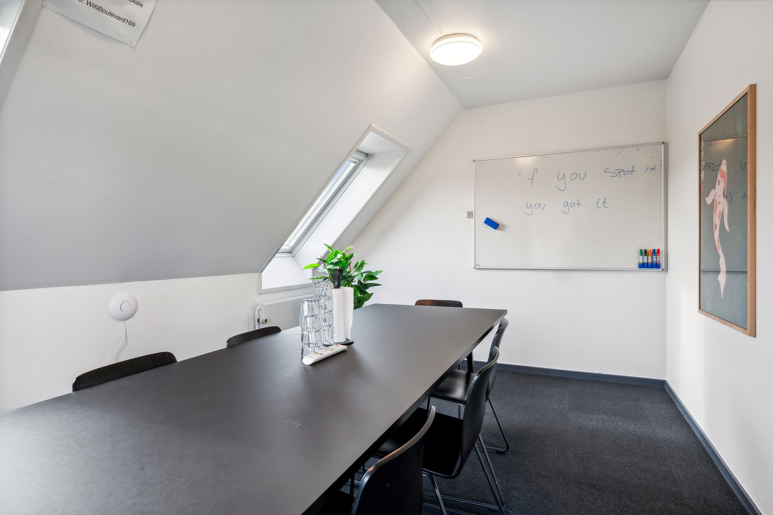 Kontorlokaler med møbler og skrivebord, hvor en skrivebordslampe er tændt, en hvid tavle er monteret og en plante står på