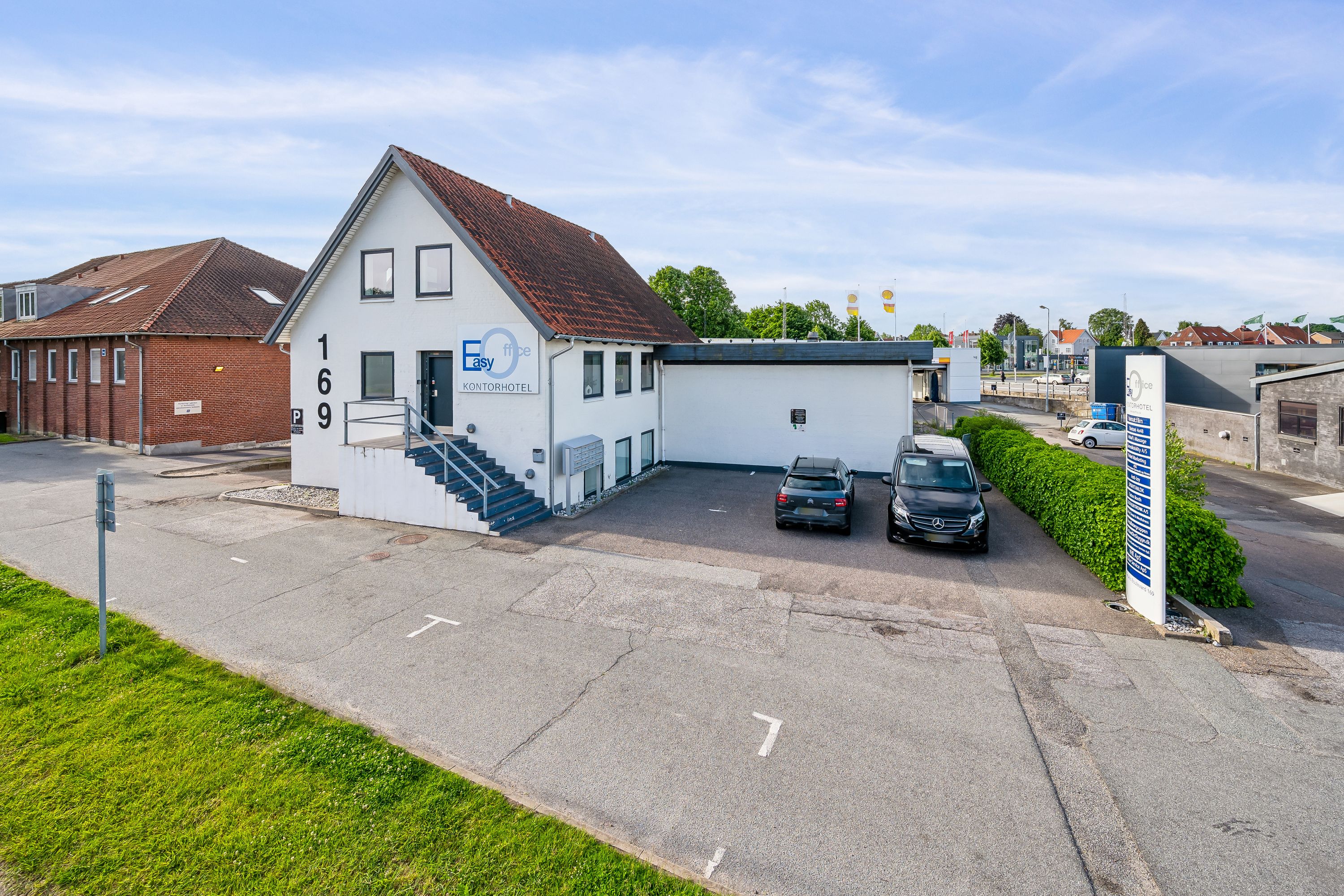 Kontorhotel med parkeringsplads og to biler, Easy Office, adresse 169, med en grøn buske og en stående søjle.
