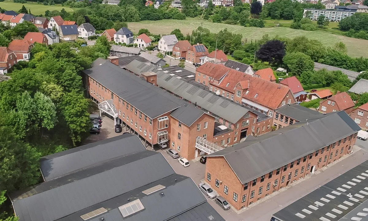 Et stort byggeanlæg med rød mursten og solceller på taget, parkeringsplads med biler og busser samt grønne træer og en bakke