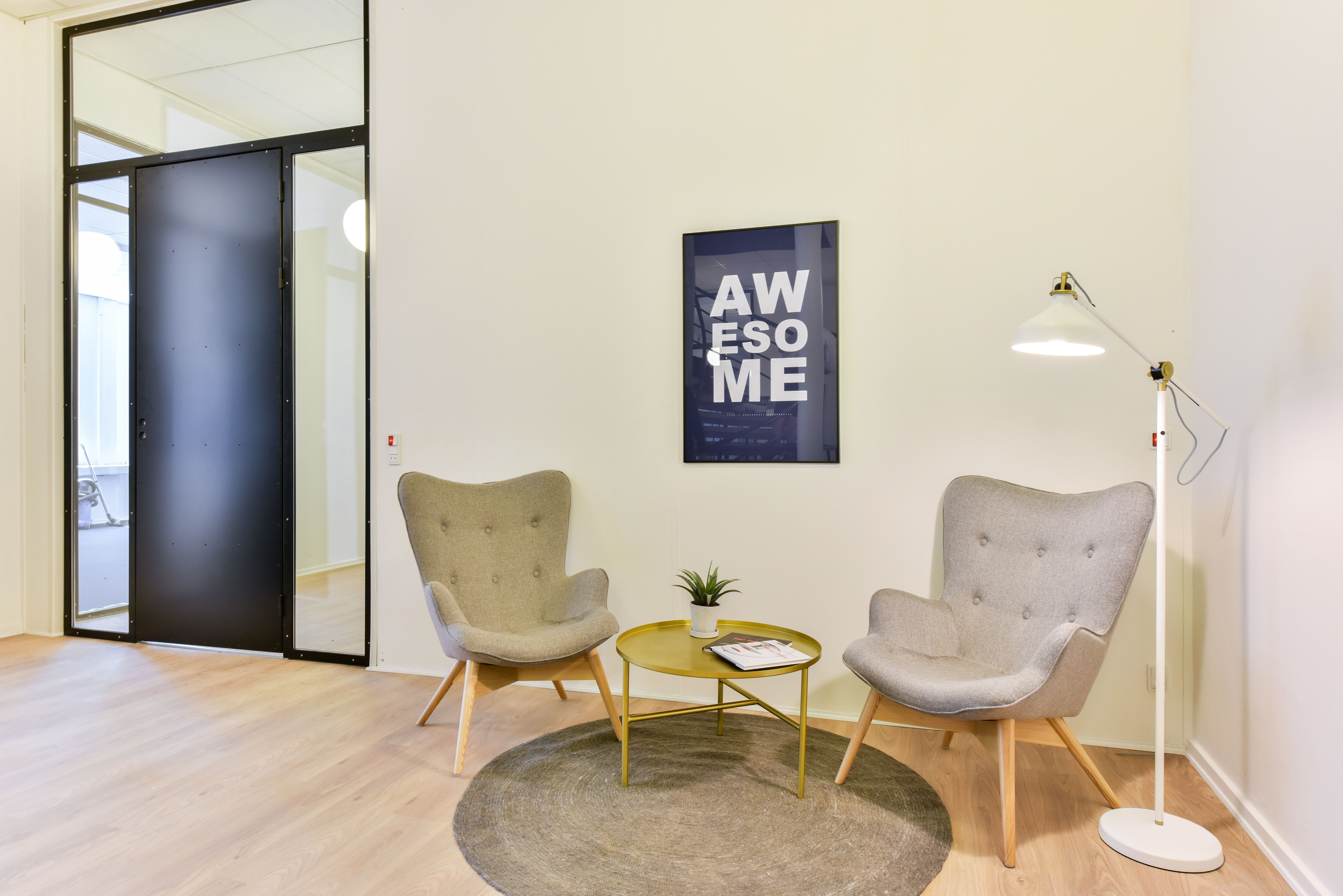 Kontorrum med to lyse lænestol, gult bord med plante og magasiner, hvid lampe og et plakat med teksten Awesome.