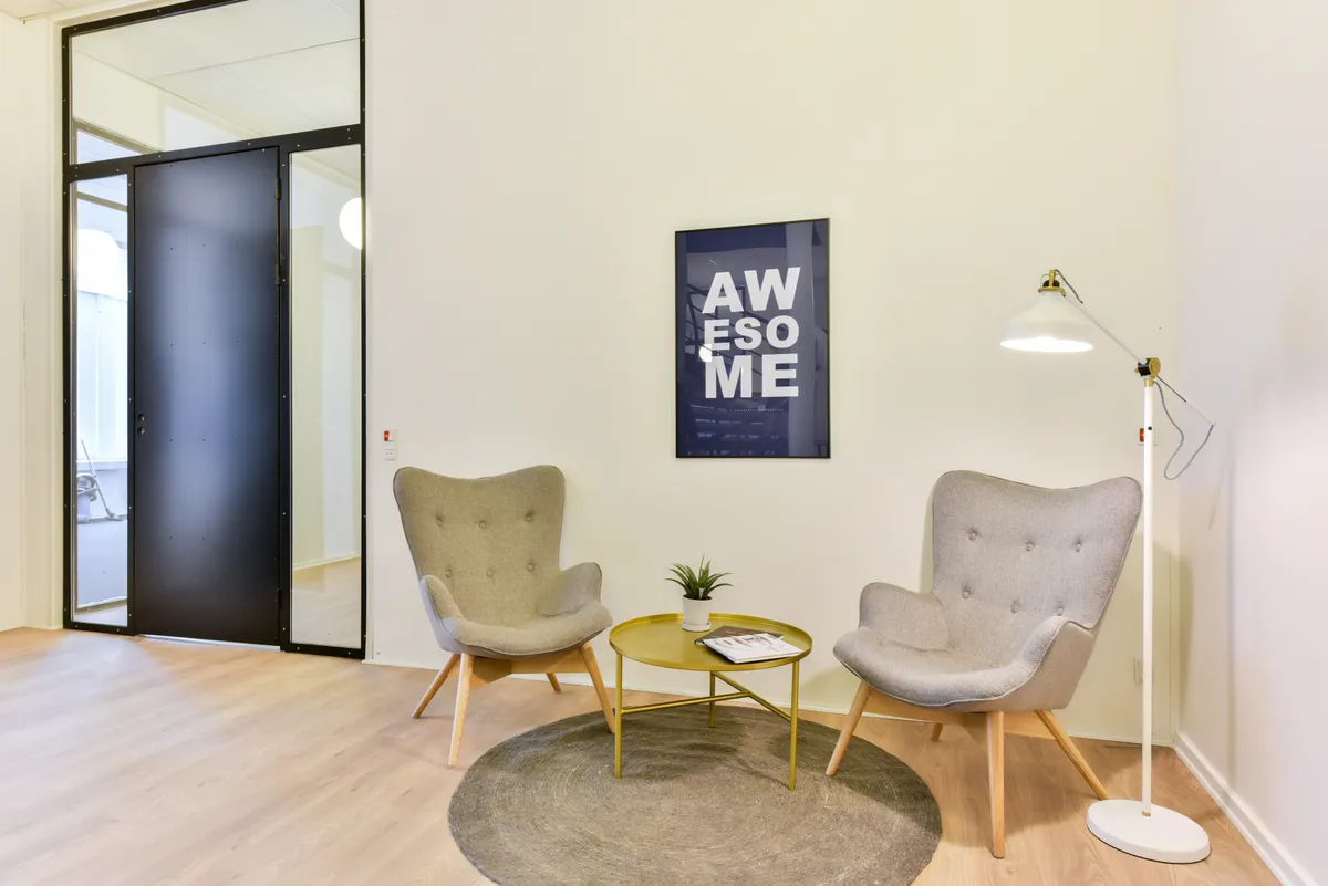 Kontorrum med to lyse lænestol, gult bord med plante og magasiner, hvid lampe og et plakat med teksten Awesome.