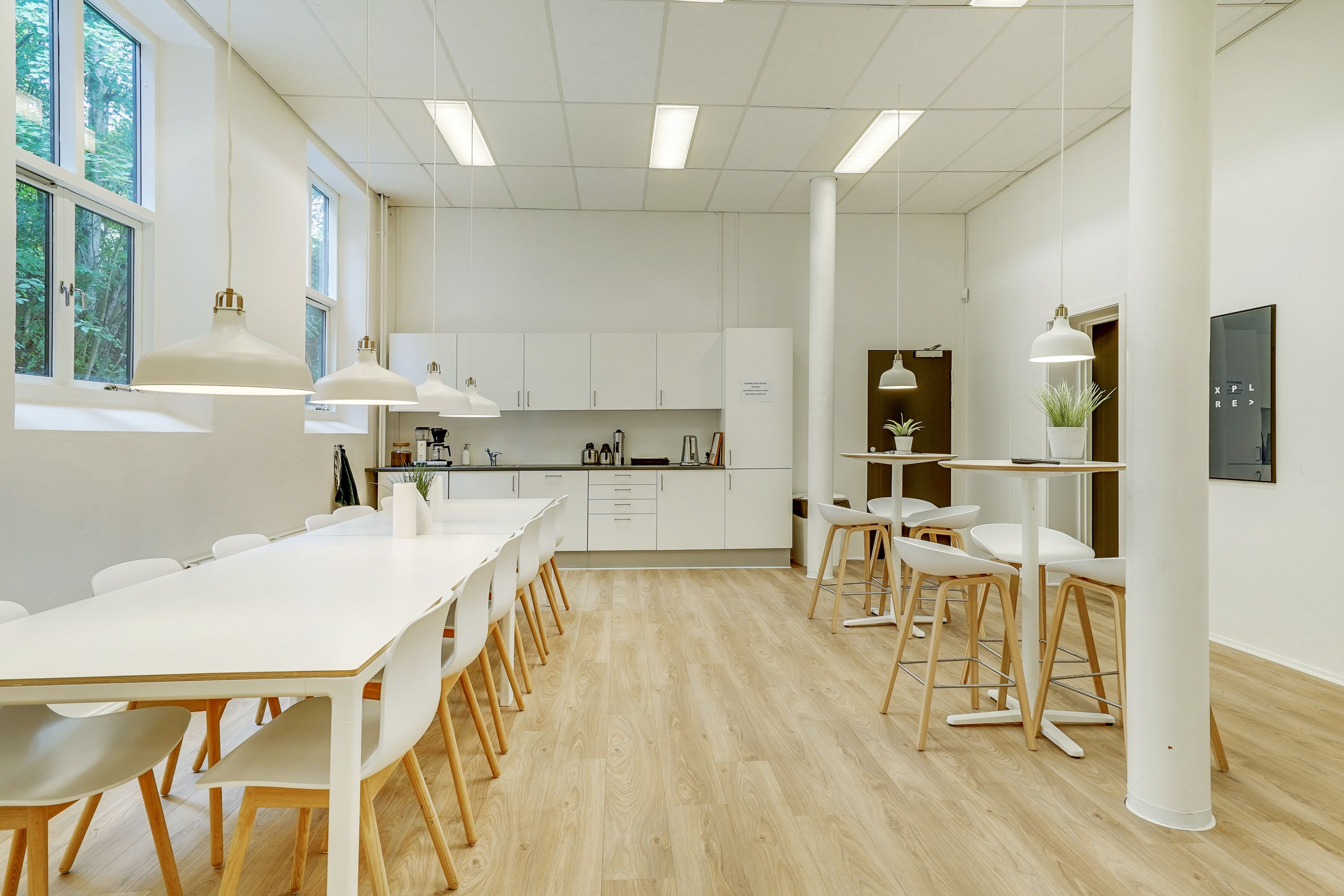Kontorrum med lange hvide borde og stole samt køkken med apparater og urtepotter på hylderne