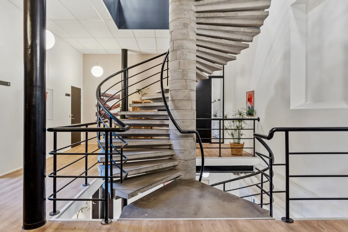 Et moderne kontorfællesskab med en spiralformet trappe og møbler i sort og grå. Der er et højt loft og en åben indgangsdør. Planter og kunstværker smykker det minimalistiske design.