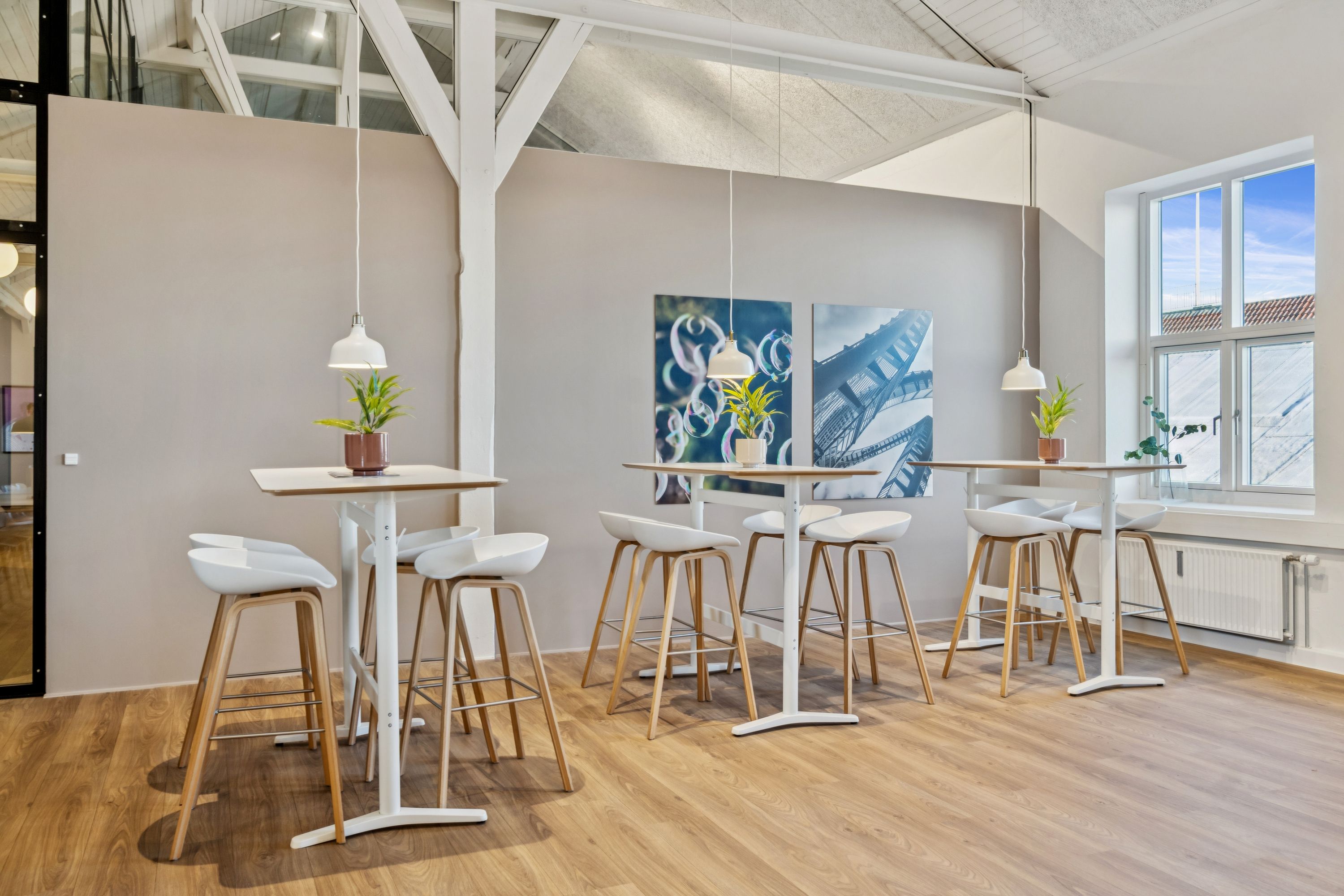 Kontorfællesskab med højt til loftet, loftsbjælker, moderne højborde og stole, lyse hængelampe, vindue med udsigt til byen og grønne planter på bordene