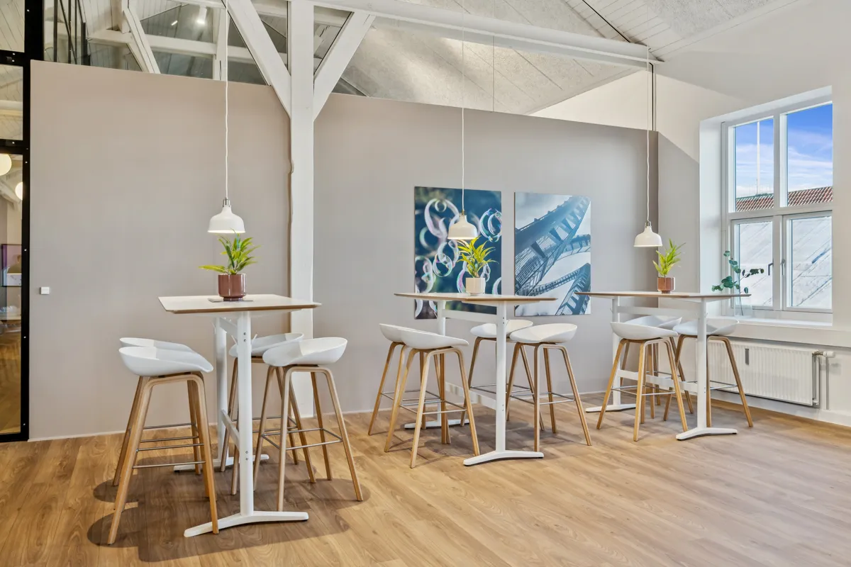 Kontorfællesskab med højt til loftet, loftsbjælker, moderne højborde og stole, lyse hængelampe, vindue med udsigt til byen og grønne planter på bordene