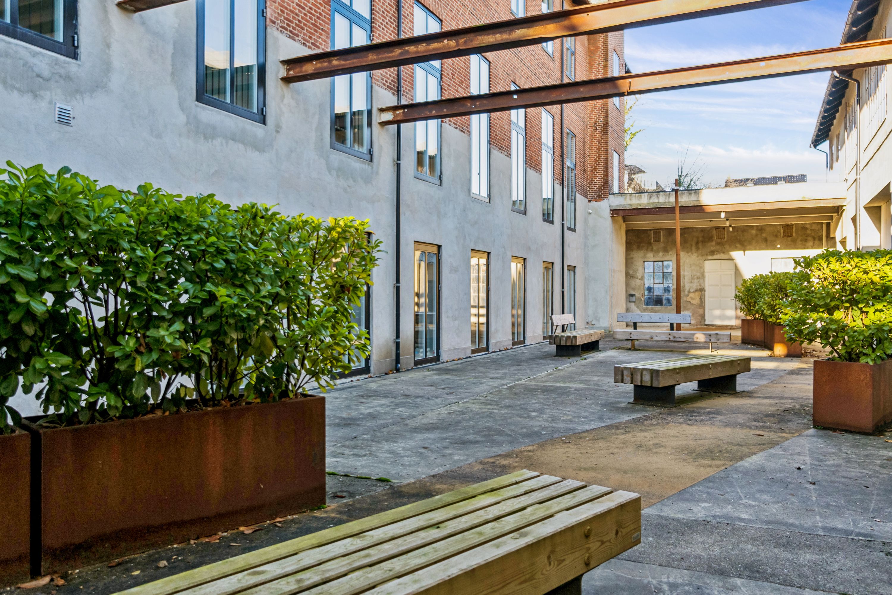Kontorfællesskab med lyse stuer og moderne udformning, rummelige møderum, parkerede biler og terrasse med grønne planter og træbænke.