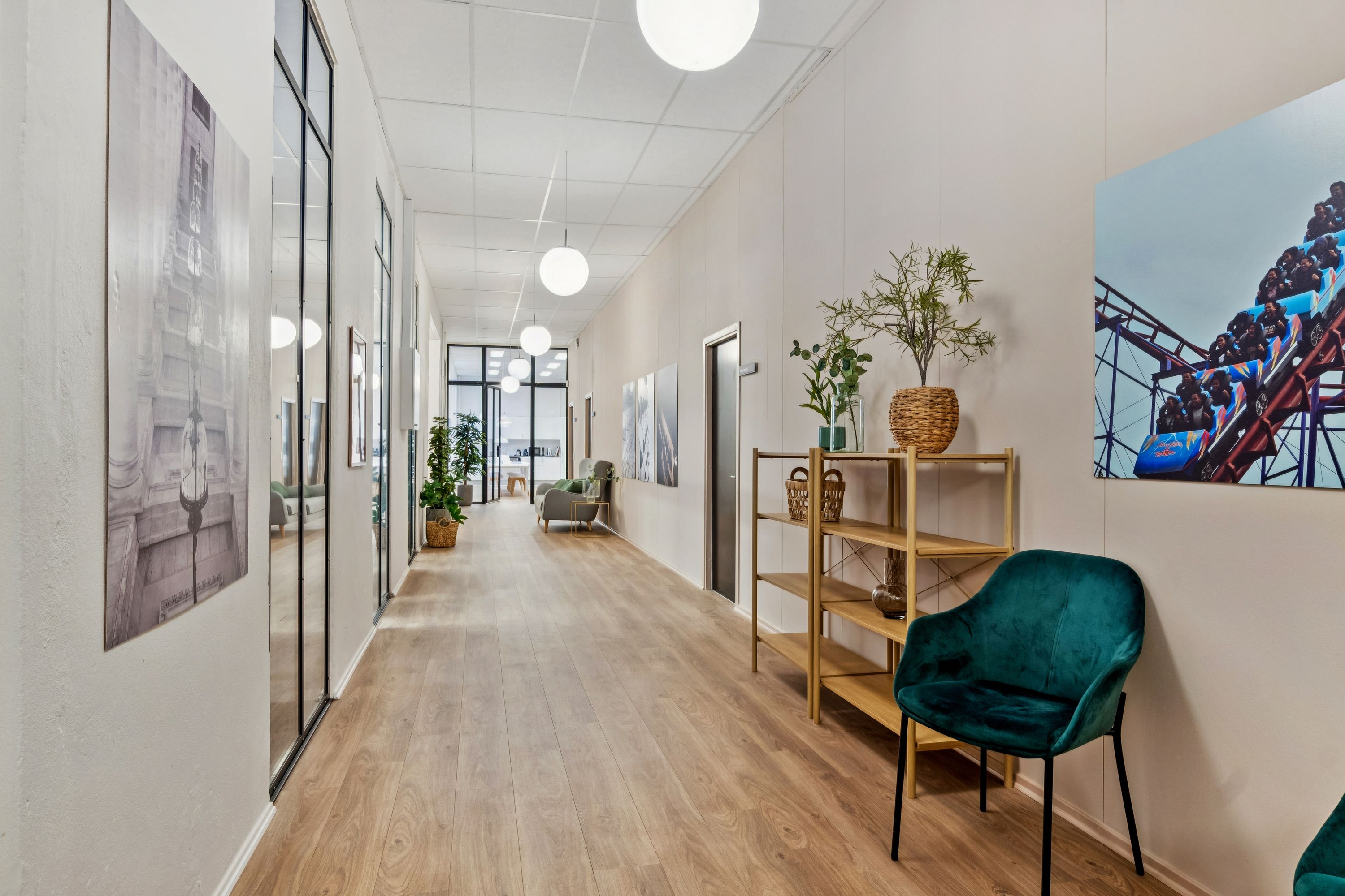 En indgangshal med trægulv og hvide vægge, to grønne stole, et højt bord med plante, et maleri af en forlystelsesby og lysekroner på loftet