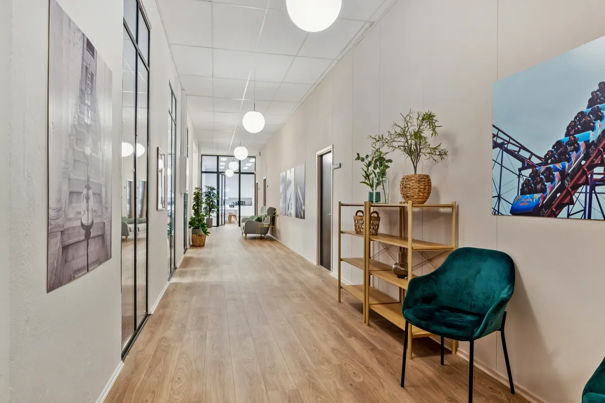 En indgangshal med trægulv og hvide vægge, to grønne stole, et højt bord med plante, et maleri af en forlystelsesby og lysekroner på loftet