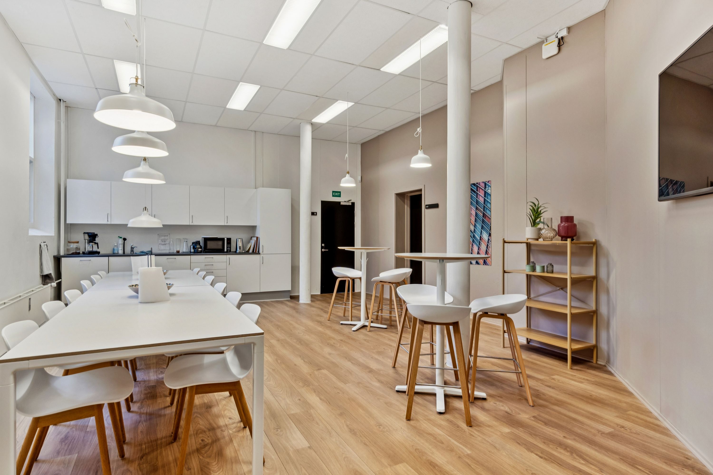 Kontorfællesskab med højloftet, hvide loftslampe, lyse loftslamper, hvide stolpe og højde. Langt hvidt bord og stole, køkken med kaffemaskine, mikroovn og køleskab.