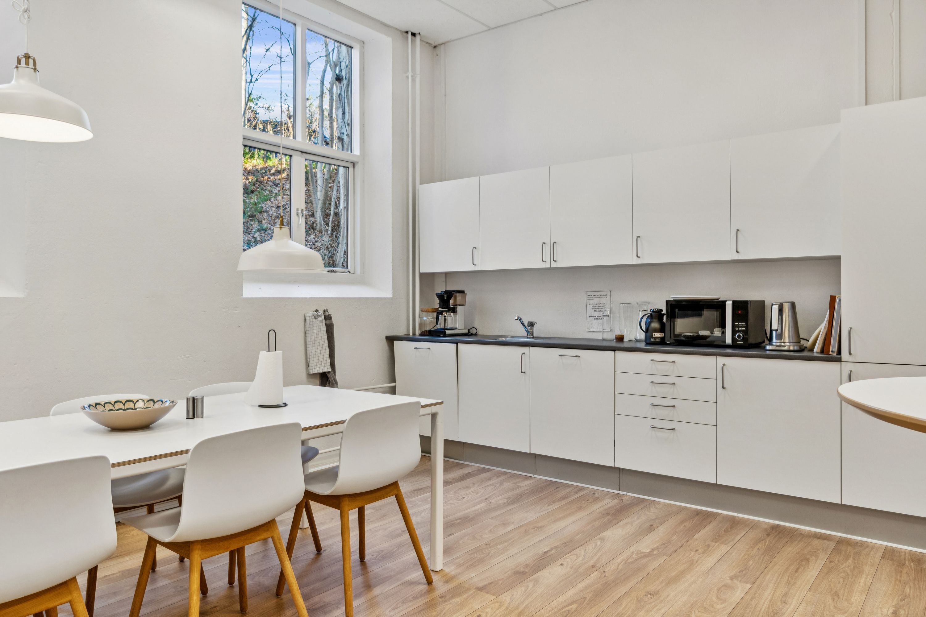 Værelse med hvide vægge og højt loft med to hængelampe. Bord og stole med massivt træ og lysegul gulv. Højt kvalitetsmøbleret køkken med hvide skabe og lysegrå bordplade.