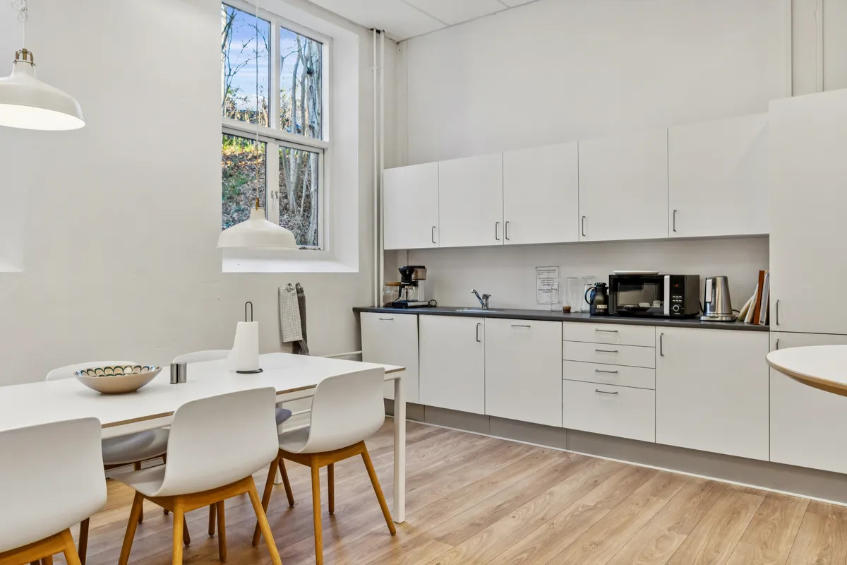 Værelse med hvide vægge og højt loft med to hængelampe. Bord og stole med massivt træ og lysegul gulv. Højt kvalitetsmøbleret køkken med hvide skabe og lysegrå bordplade.