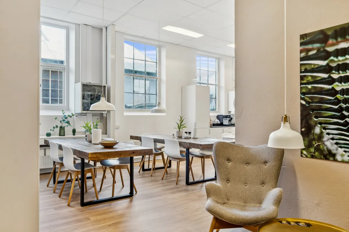 Kontorfællesskab med to store borde, hvide stole, lysekroner og et maleri på væggen samt en sofa med et bord i guldet.