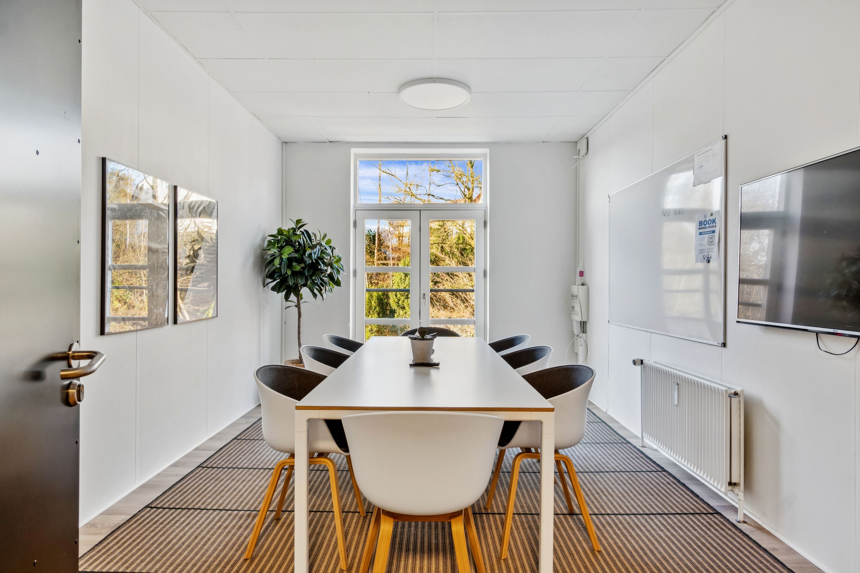 Kontorrum med hvide vægge og bord med stole, hvor gulvet er dækket af mønstret gulvtæppe og skov i baggrunden.