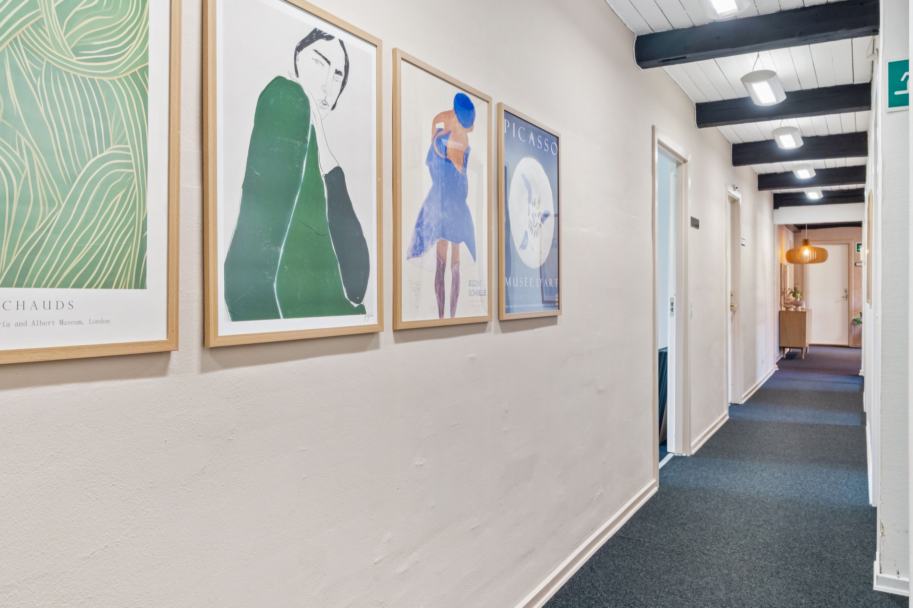 Kunstværker hænger på væggen og lyser op med lysekroner. Væggene er hvide og gulvet er lyseblå. Gangen er lyseblå med lysekroner og en åben dør.