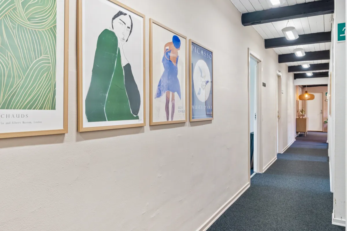 Kunstværker hænger på væggen og lyser op med lysekroner. Væggene er hvide og gulvet er lyseblå. Gangen er lyseblå med lysekroner og en åben dør.