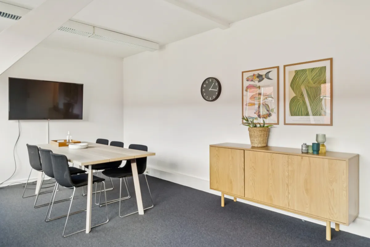 Et moderne kontorlokal med en stor hvid væg og et stort skærmbillede på væggen. Der er en høj kvalitetsbord med stolpe og en plante i kurven.