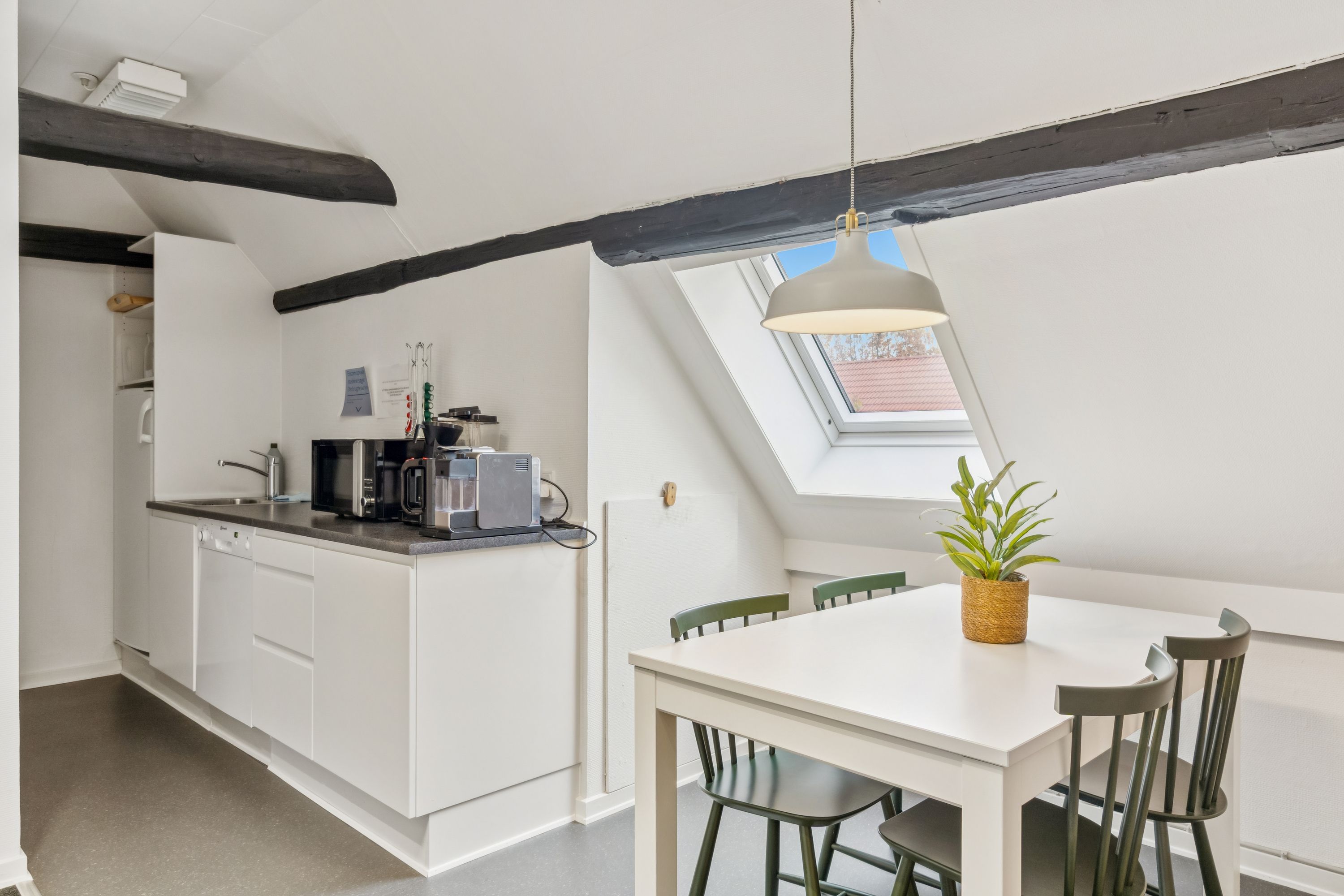 Et lille køkken med lyse loftsbjælker, et hvidt bord og lyse grønne stole, en plante i kurven og en loftsvindue med udsigt til en rødt tag