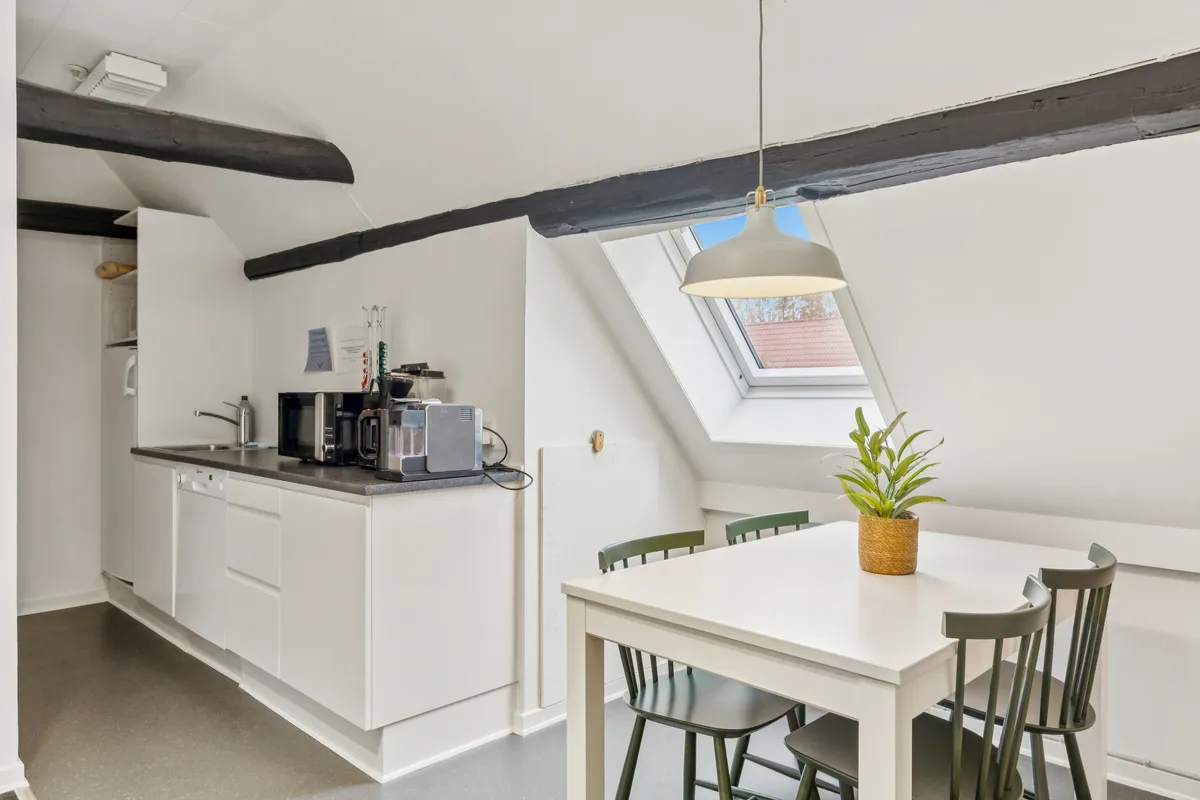Et lille køkken med lyse loftsbjælker, et hvidt bord og lyse grønne stole, en plante i kurven og en loftsvindue med udsigt til en rødt tag
