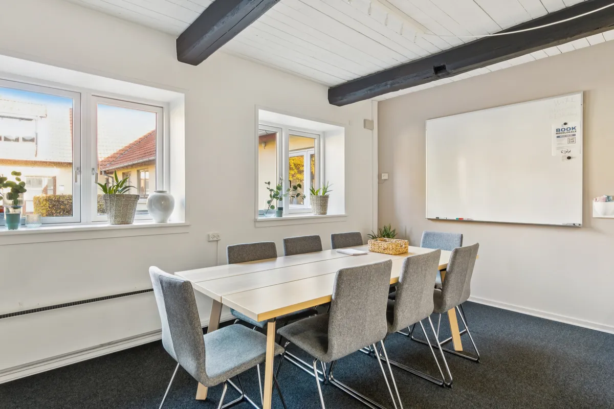 Kontorrum med en lang bord og otte stole rundt om bordet samt tre vinduer med potter og plantevægge