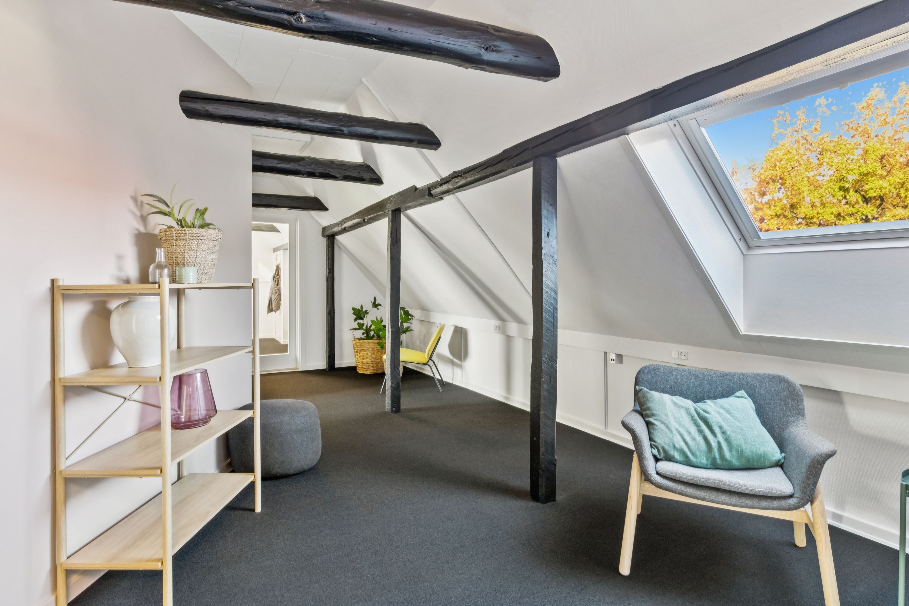Et moderne loftsværelse med lyse loftbjælker og et komfortabelt sæt med stol og pude samt et hyggeligt plantearrangement på hylden og en udsigt til træerne udenfor