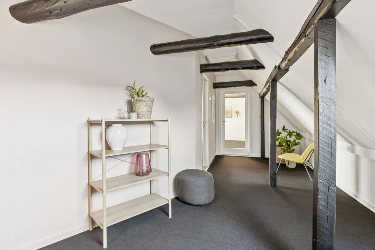 Alt-tekst: Loftsværelse med højloft og træbjælker, hvid væg, gulv og gulvtæppe, høj hylde og møbler, inkl. plante og lysestager