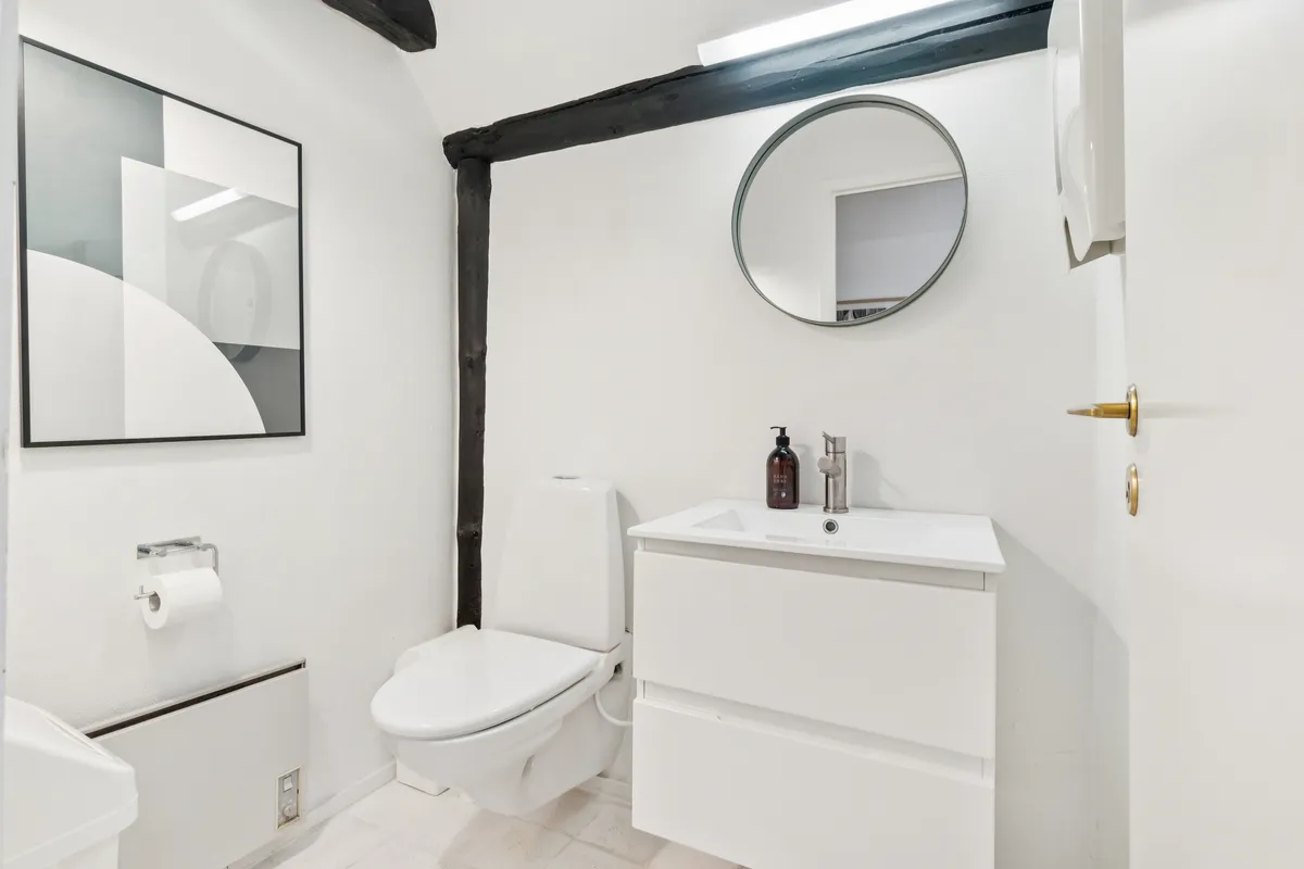 Badrum med hvidt toilet, hvidt spejl, hvidt vandhaner, hvidt vask og hvid væg. Toiletpapir holder og elektrisk radiator på væggen