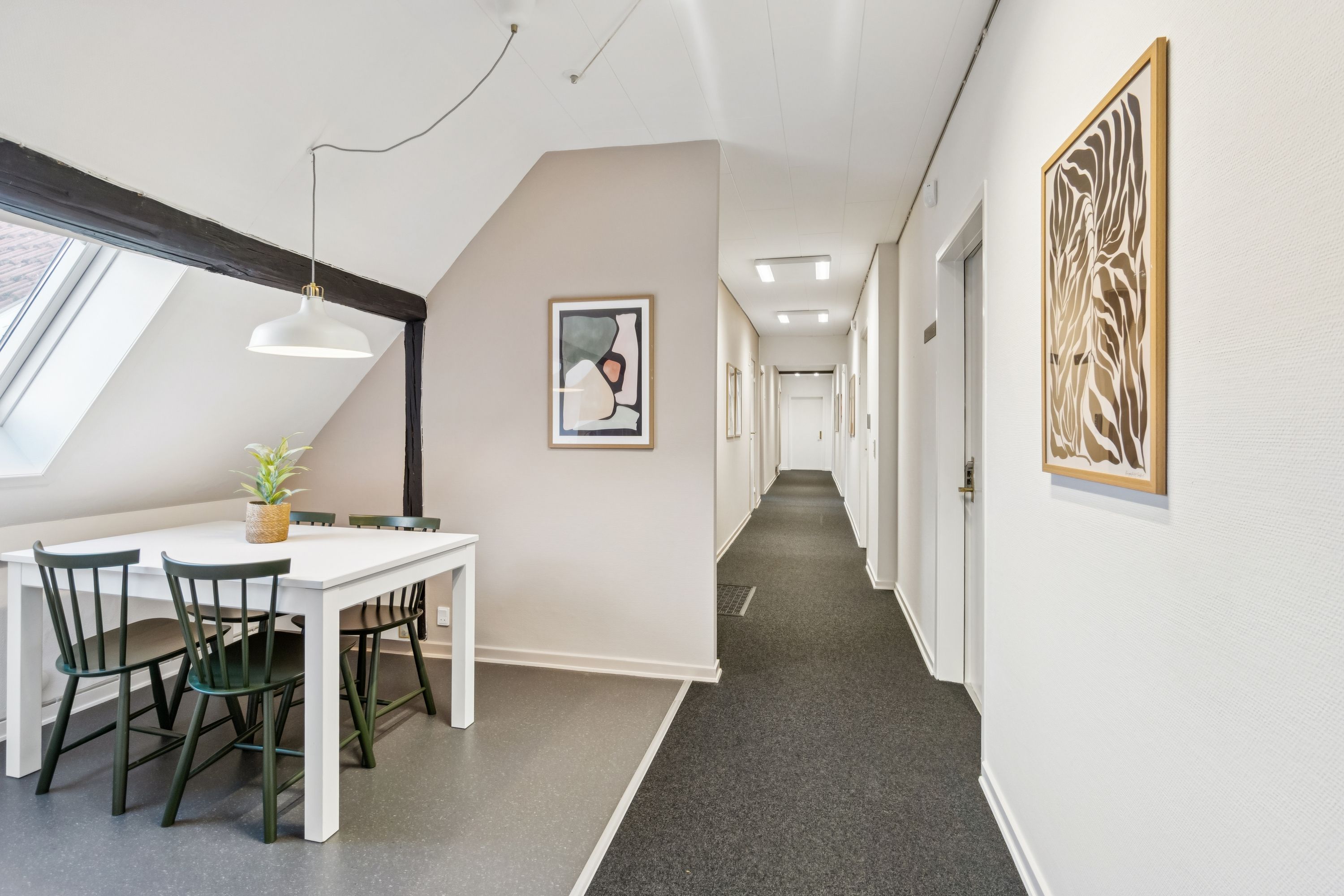 En kontorfællesskab med et hvidt spisebord og grønne stole samt en plante og loftsvinduer med naturlys