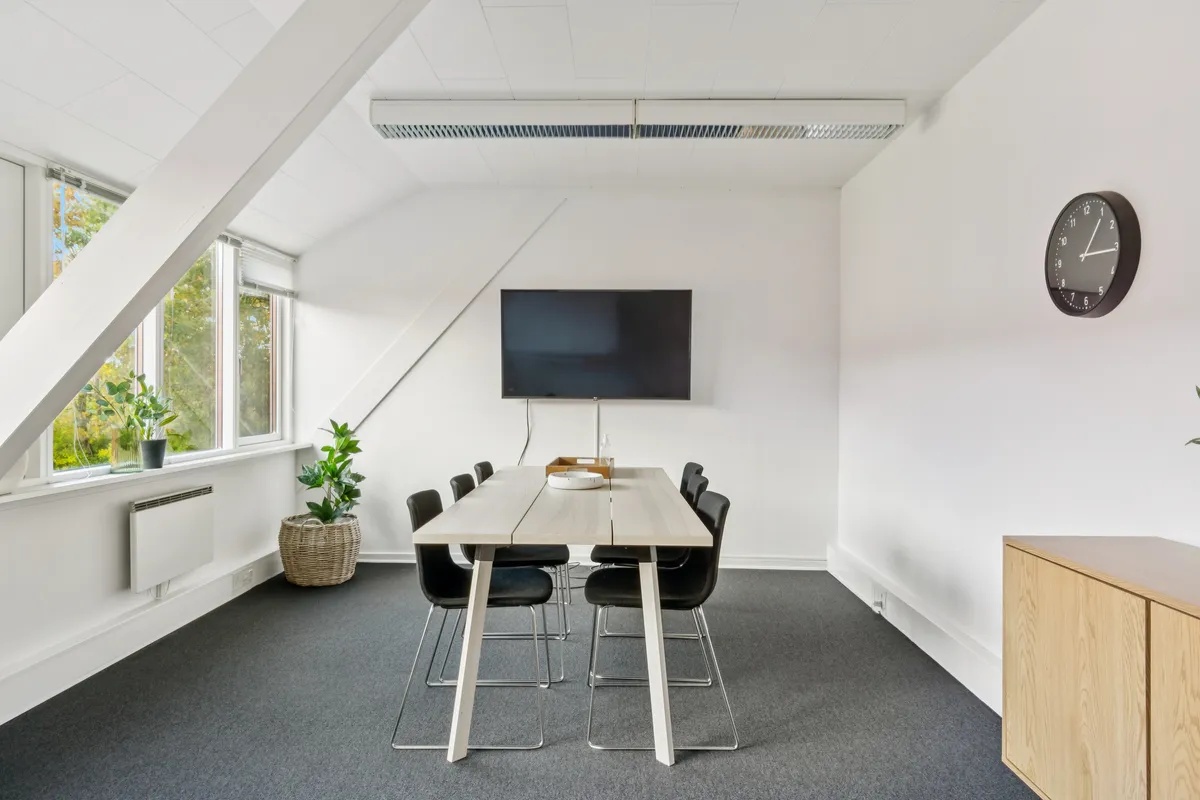 Kontor med højt loft, hvide vægge, møbler og plantepot. Sort ur, tv og vinduer med udsigt til grønne træer