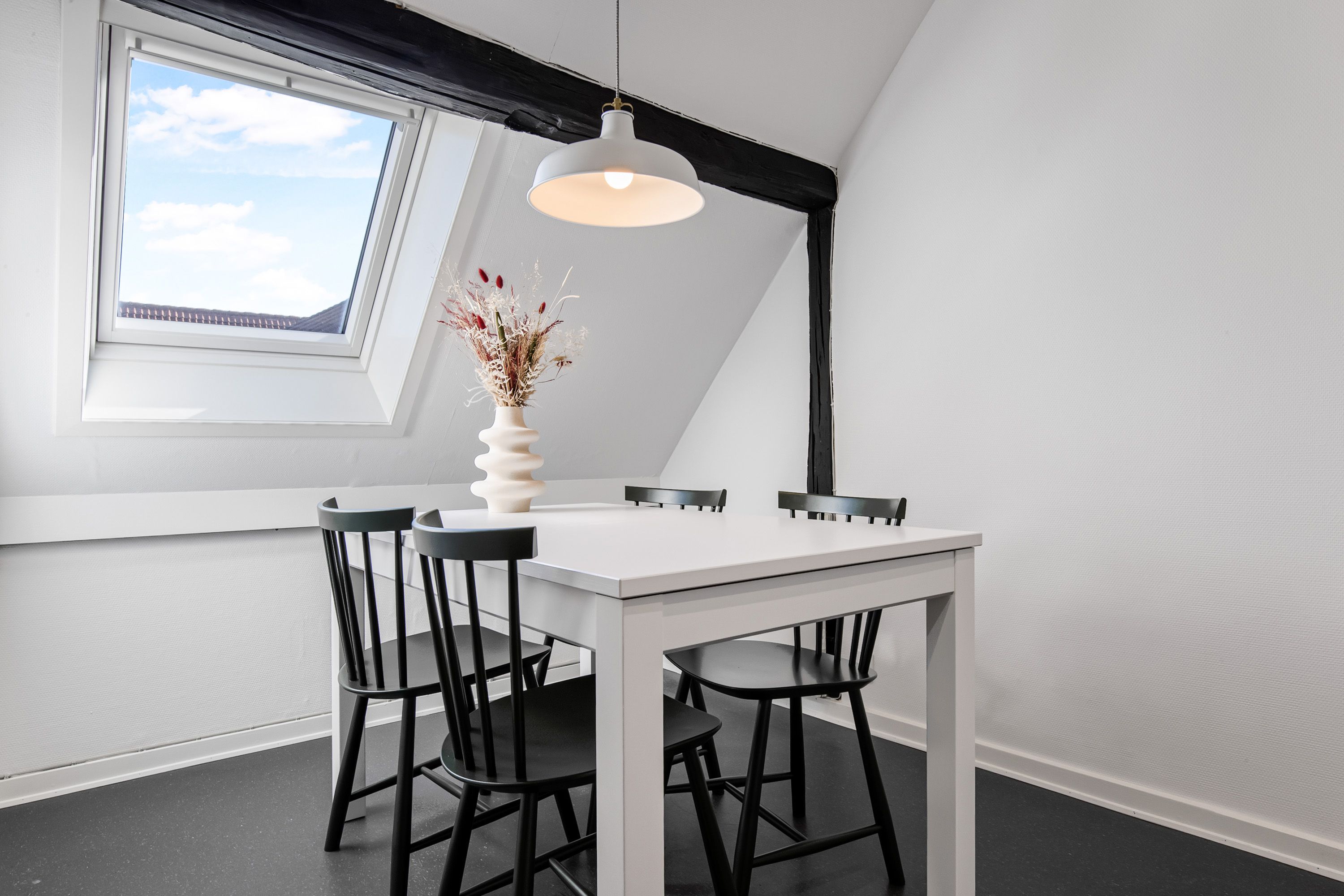 En hvid spiseplads med et hvidt bord og fire sort bordstolpe. Et hvidt loftsvindue og et hængende lampe med lyseperler. Et