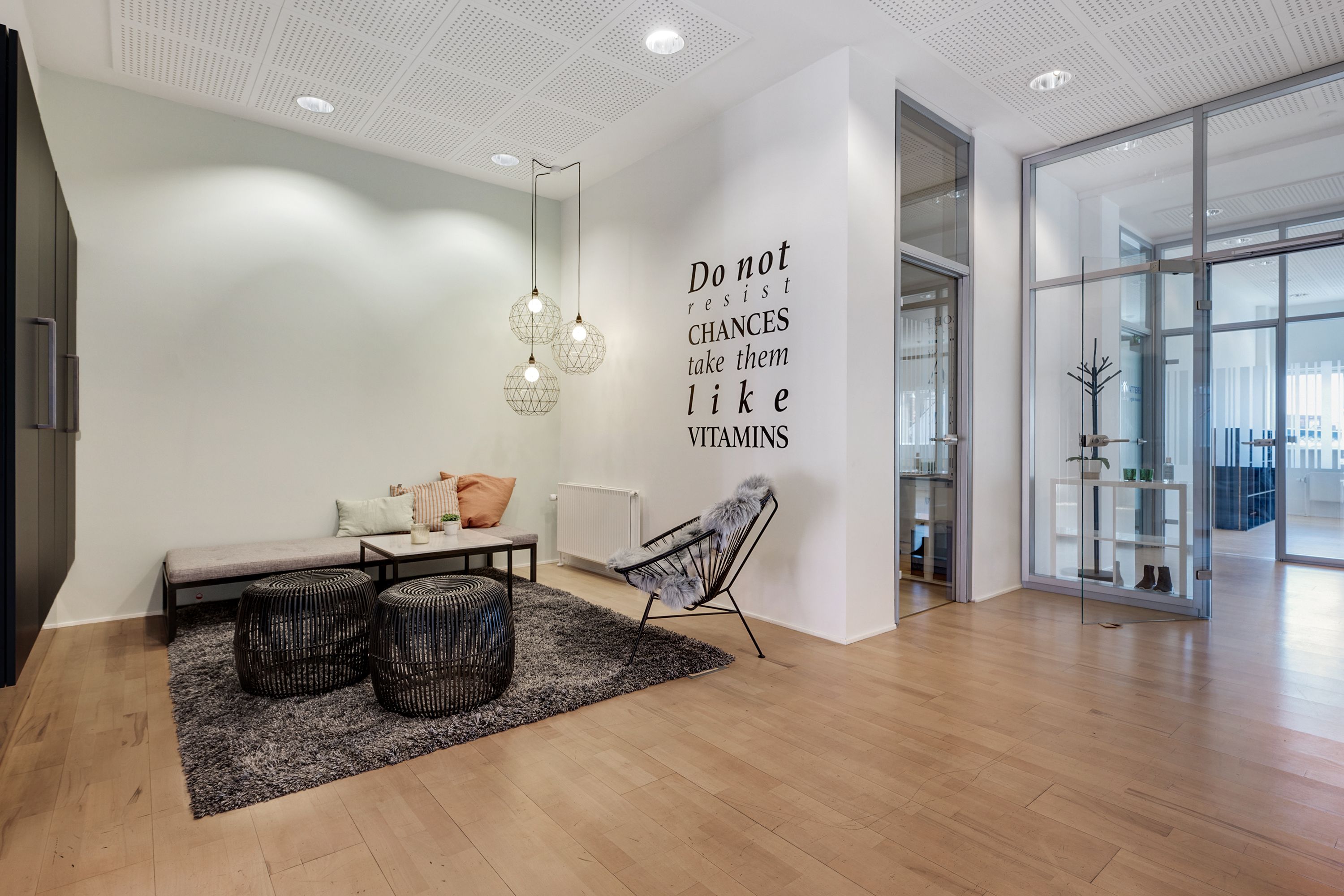 Indretning med højloftede lokaler og møbler, der inkluderer en sofa, stole og bord, samt en væg med citat og plante.