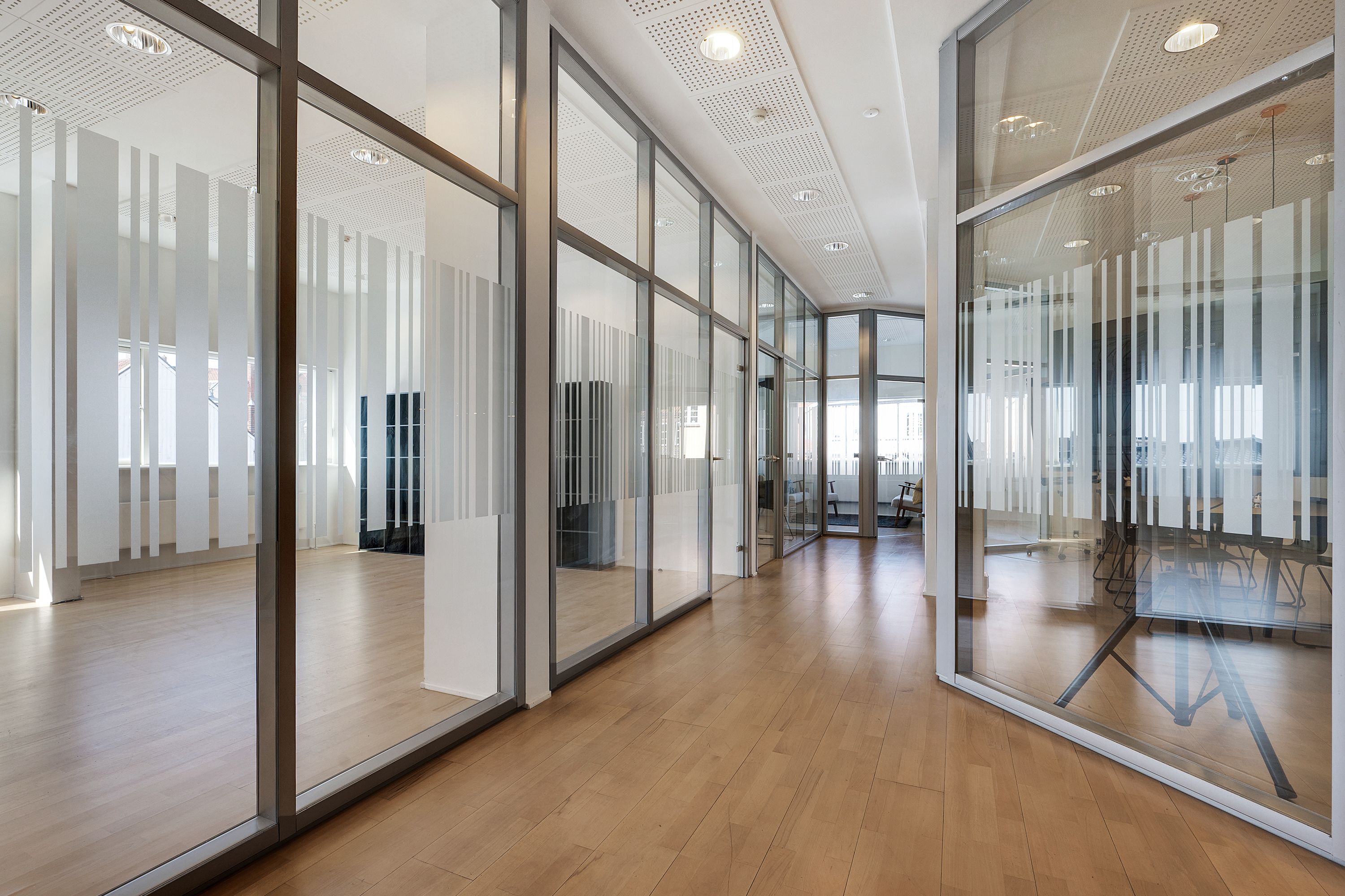 Kontorområde med glasvægge og trægulve. Glasmure og spejle reflekterer hvide lofter og lyse loftslamper. En række stole og