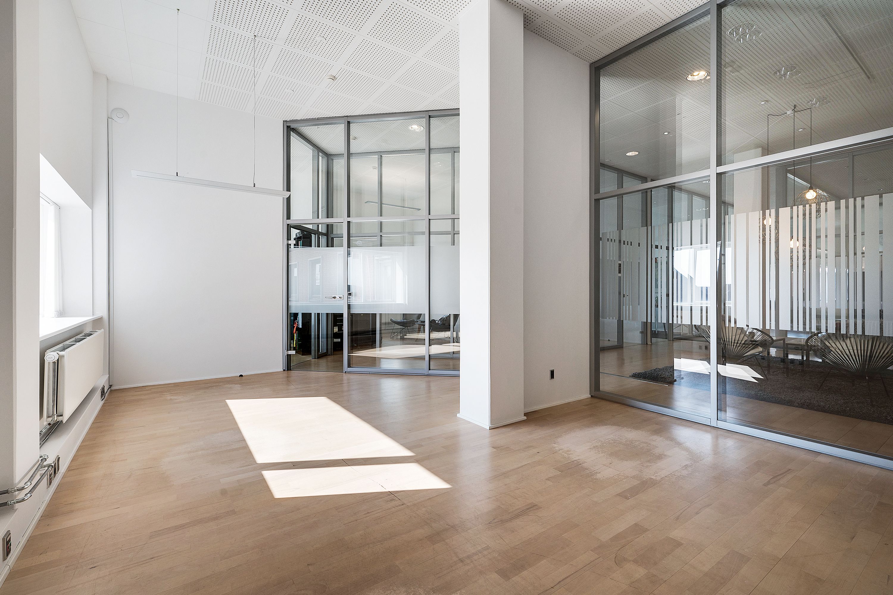 Et kontorlokale med lyse trægulve og store glaspartier. På gulvet er der mærkbare solstråler, og der er en hvid væg med et