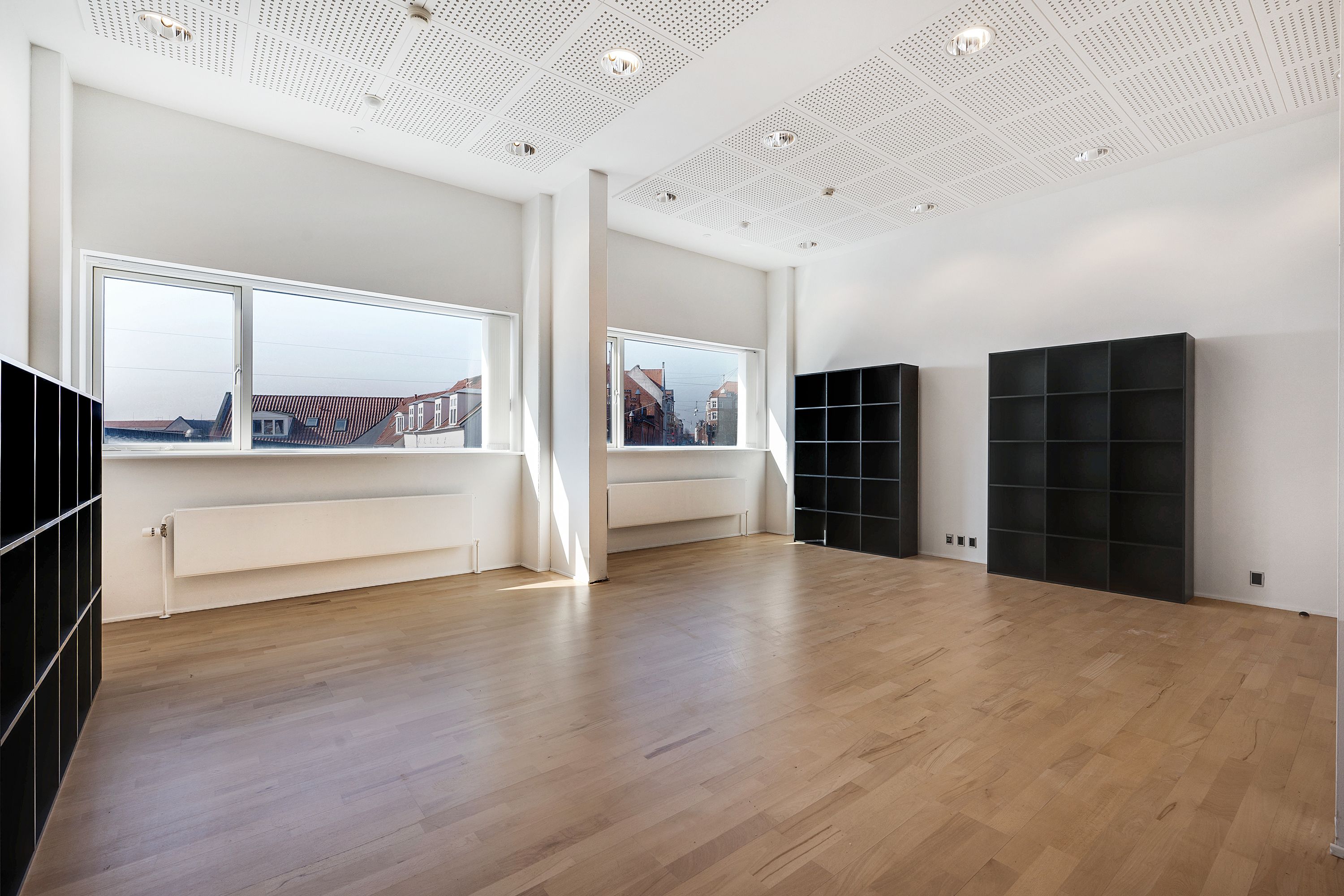 Et stort kontorlokum med lyse lofter og store vinduer og to store, sorte bogreoler på højre side og en på venstre side.