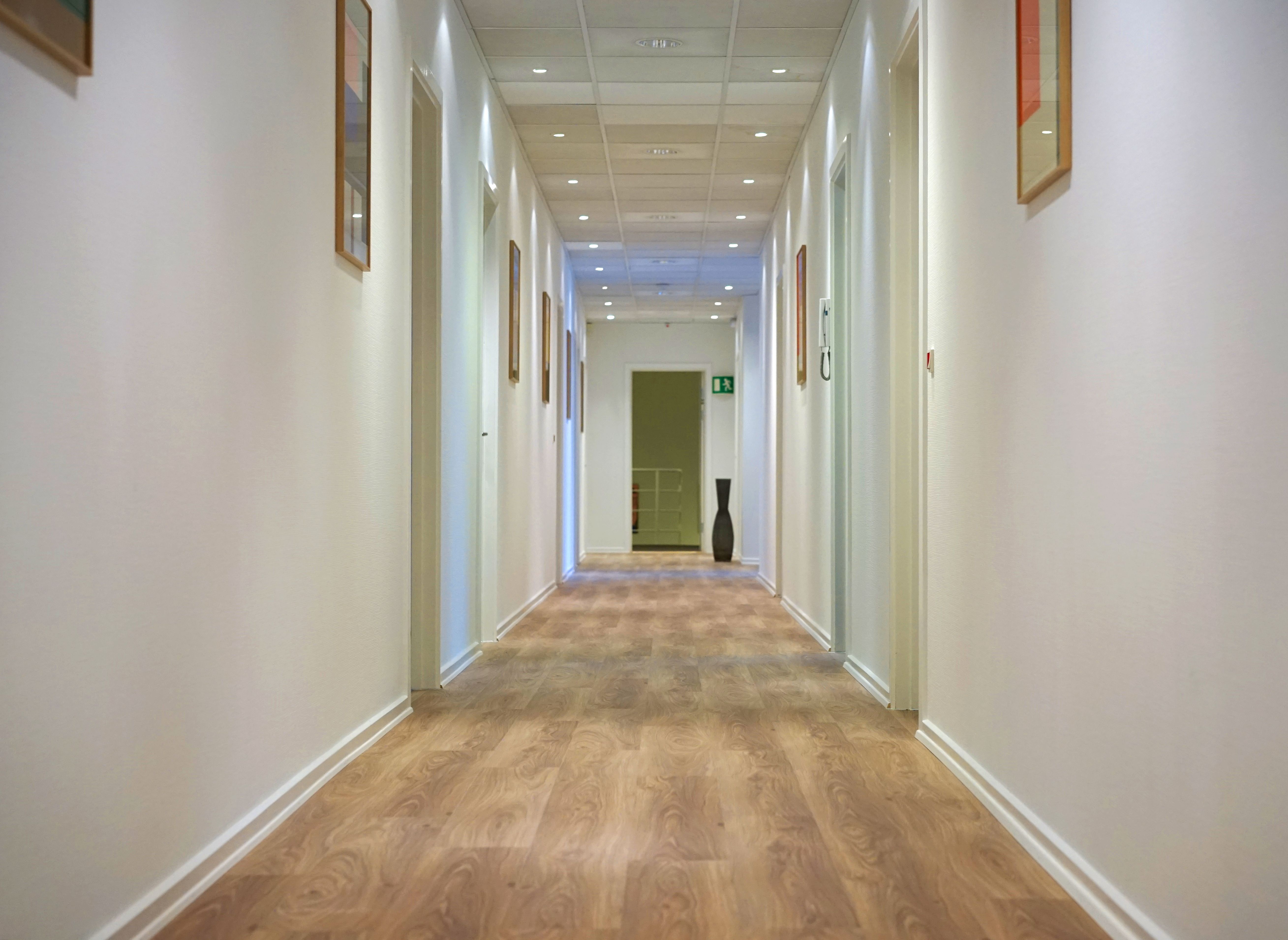 En lang korridor med hvide vægge og lyse gulvbelægning med mønstre, et stort sort glasvase og et grønt dørskildt med rødt