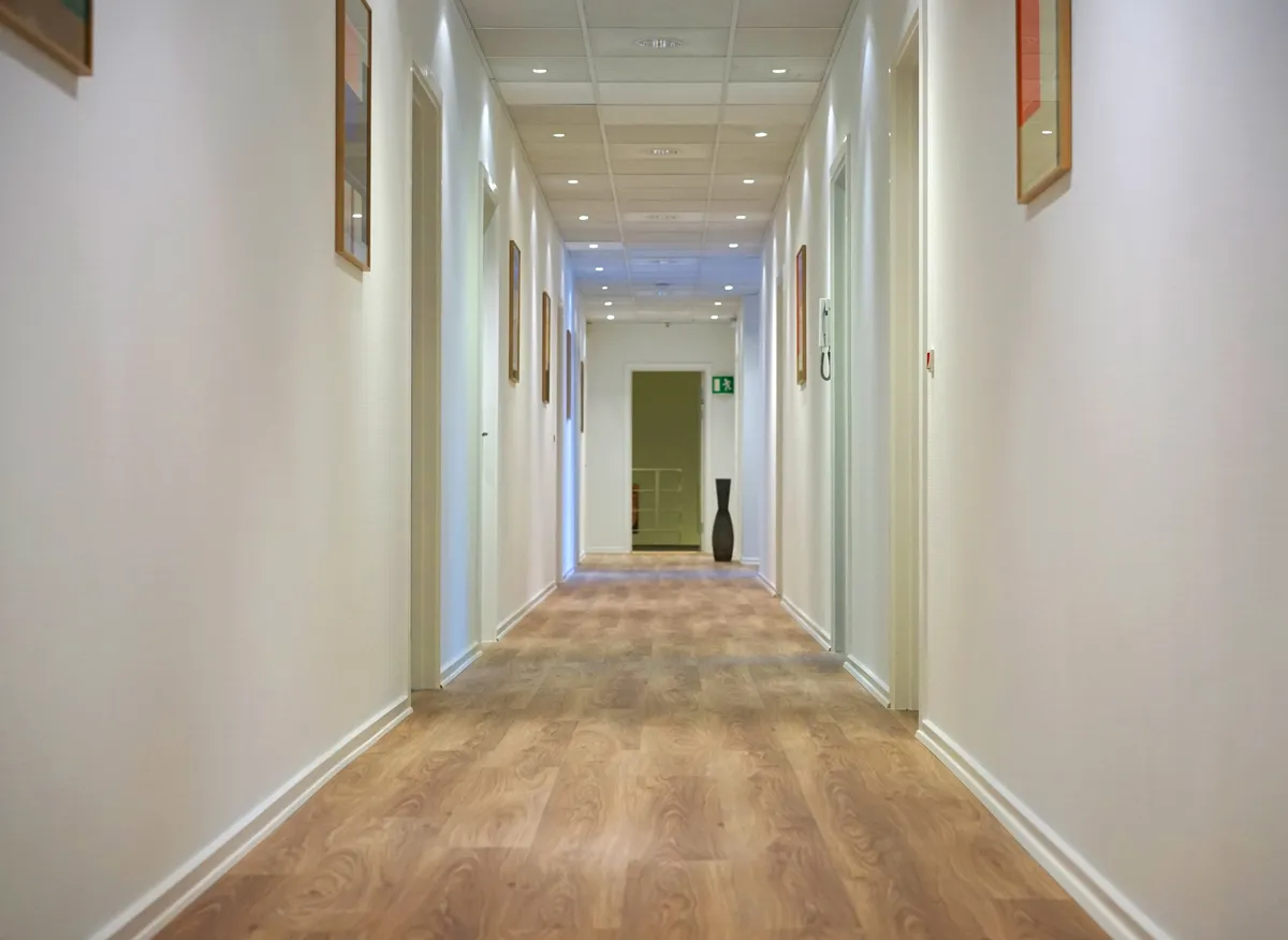 En lang korridor med hvide vægge og lyse gulvbelægning med mønstre, et stort sort glasvase og et grønt dørskildt med rødt