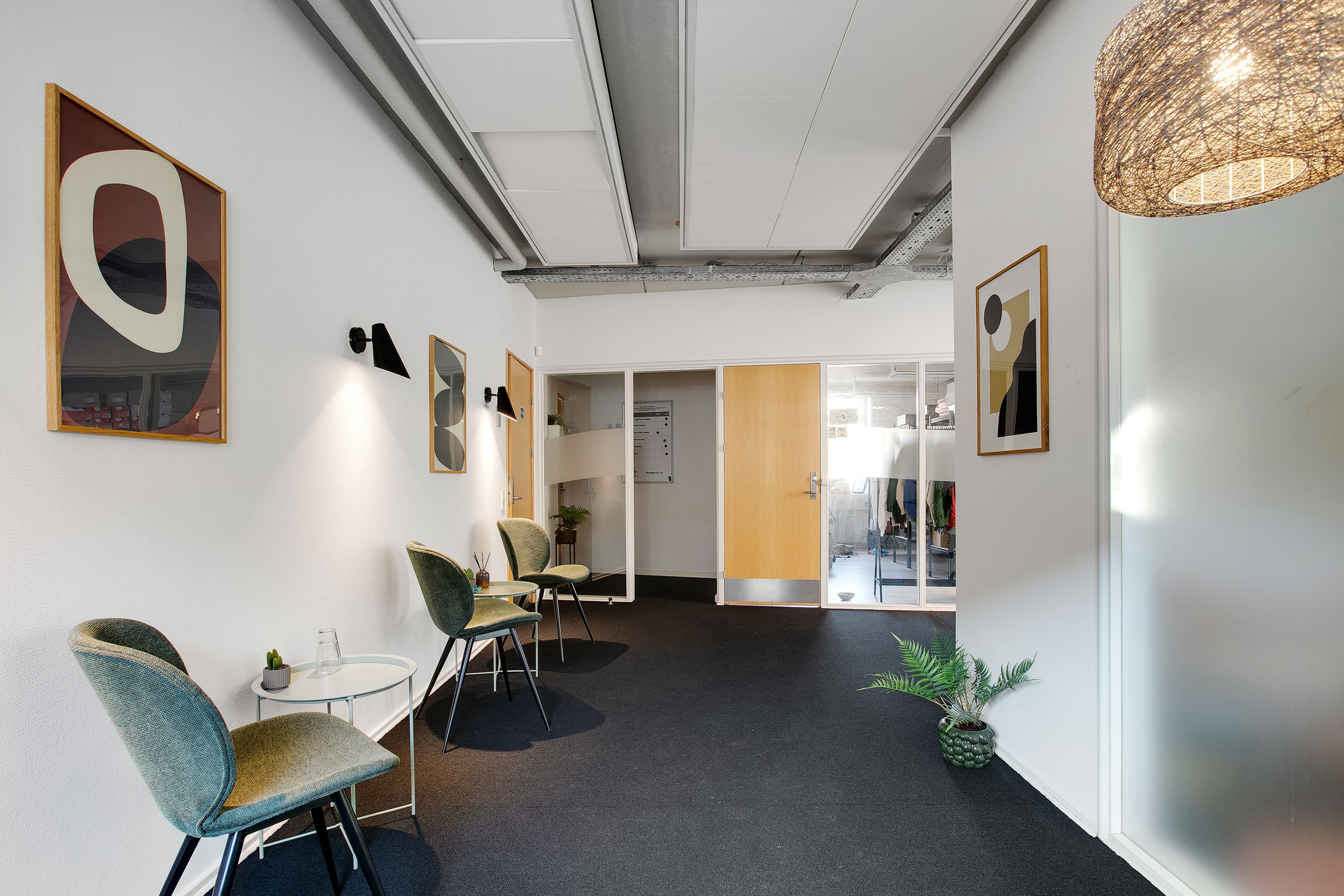 Kontorgang med lyse grønne stole og et lille bord med en skulptur og plante. Der er plakater på væggen og et lysekron.