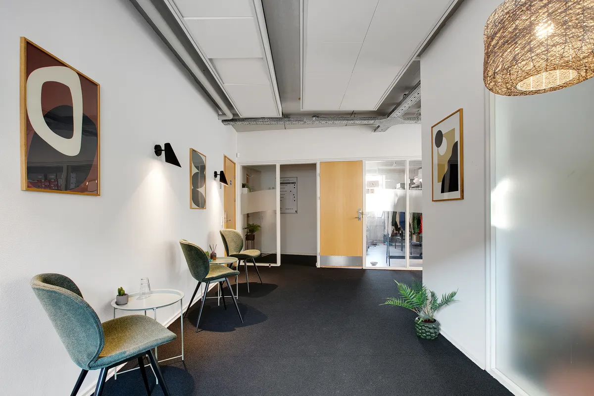 Kontorgang med lyse grønne stole og et lille bord med en skulptur og plante. Der er plakater på væggen og et lysekron.
