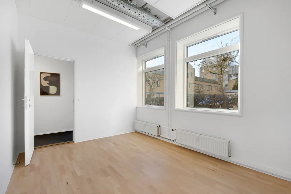 Hvidt kontorlokum med trægulv og store vinduer, lyse radiatorer og abstrakt maleri på væggen