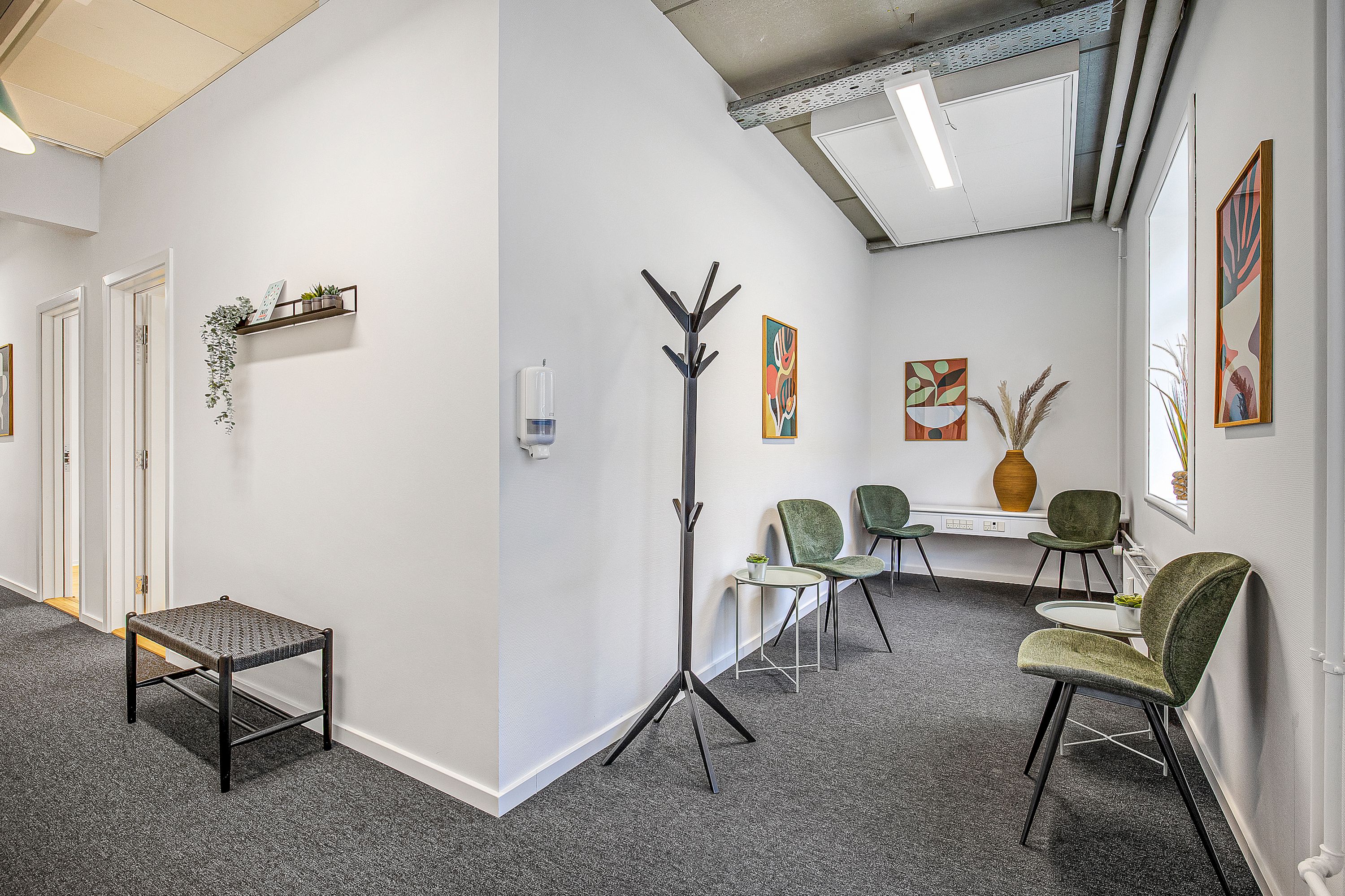 Møbelstileret kontorfællesskab med kaffeborde, grønne stole, gulvvarme, håndsæbe og loftslampe. Blomster og planteværk er spredt rundt på væggen.