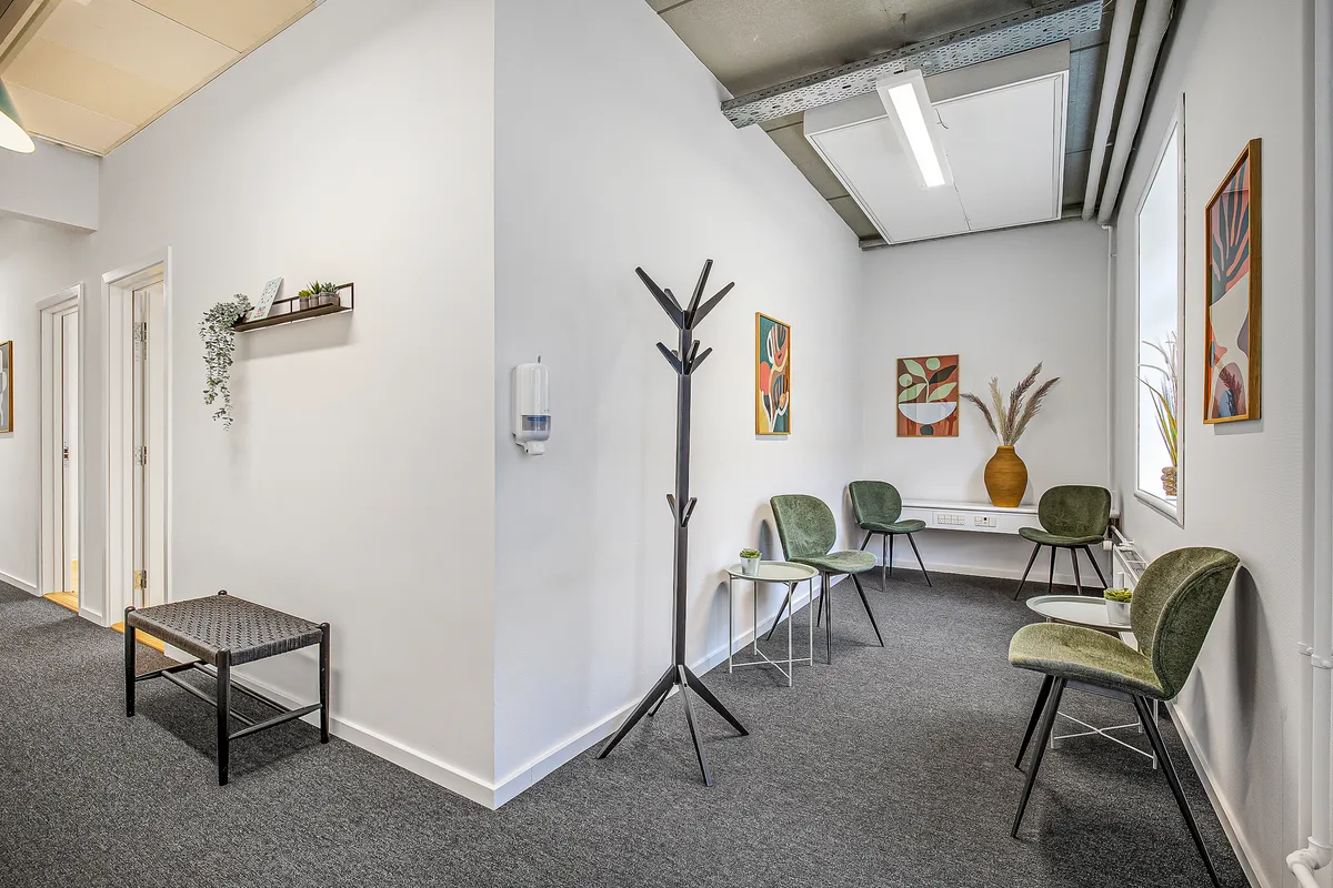 Møbelstileret kontorfællesskab med kaffeborde, grønne stole, gulvvarme, håndsæbe og loftslampe. Blomster og planteværk er spredt rundt på væggen.