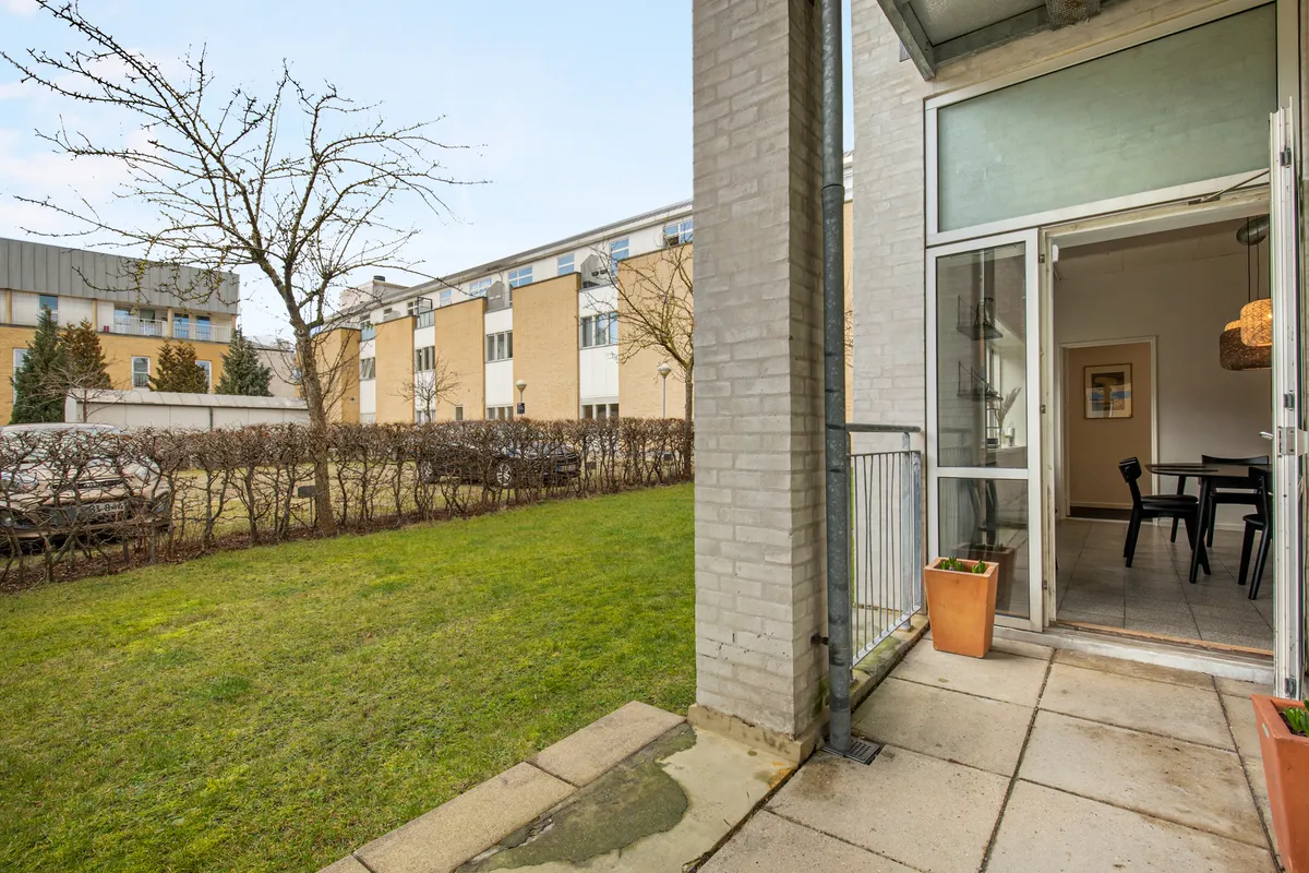 Terrasse med udsigt til græsplæne og gårdhave, åben dør til lejlighed, gulvbelægning, planter, lysekroner, træer og bygninger i baggrunden