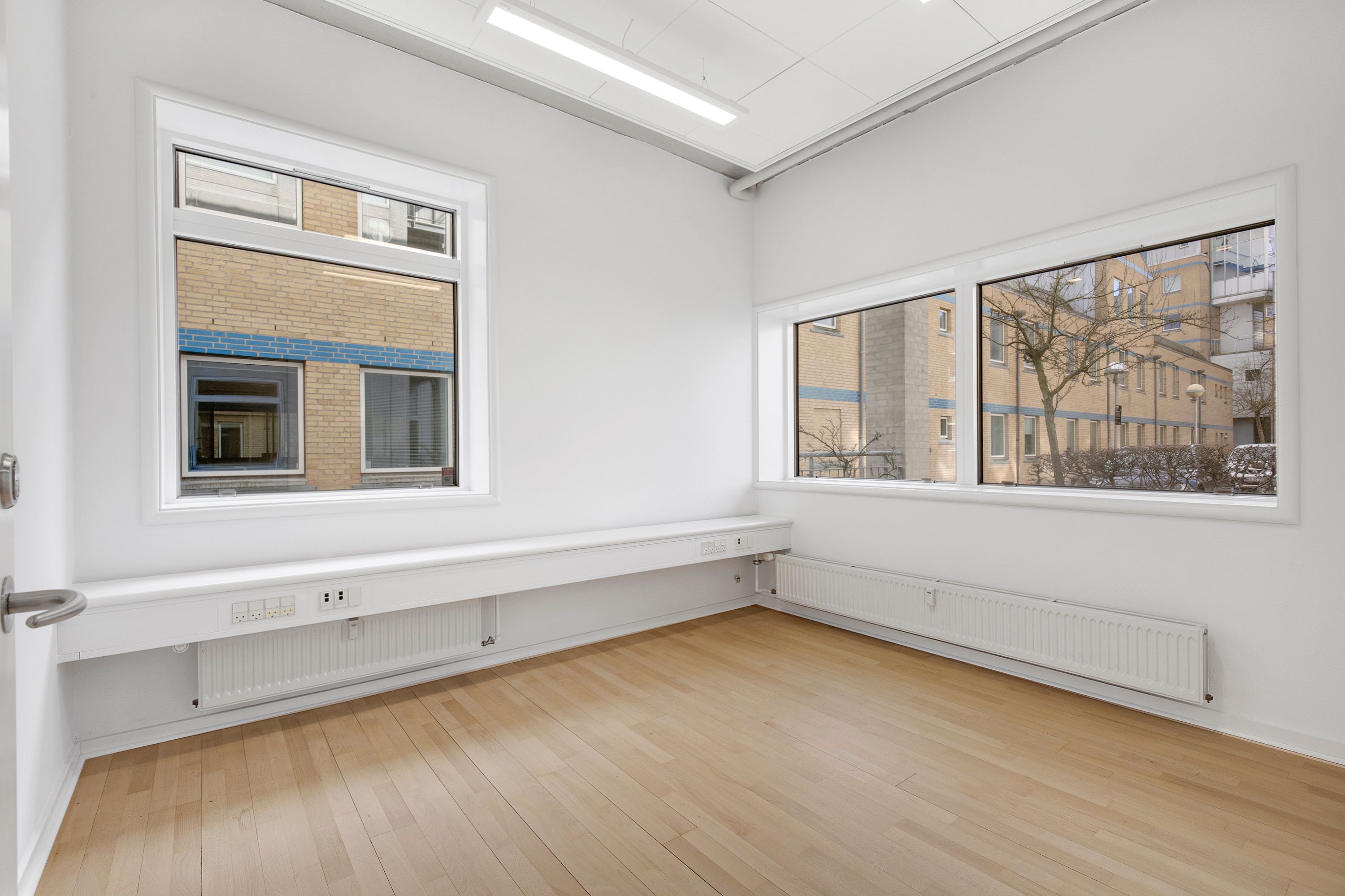 Et moderne kontor med lyse gulve, hvide vægge og store vinduer. Et hvidt radiator er monteret i væggen og en dør er åben.