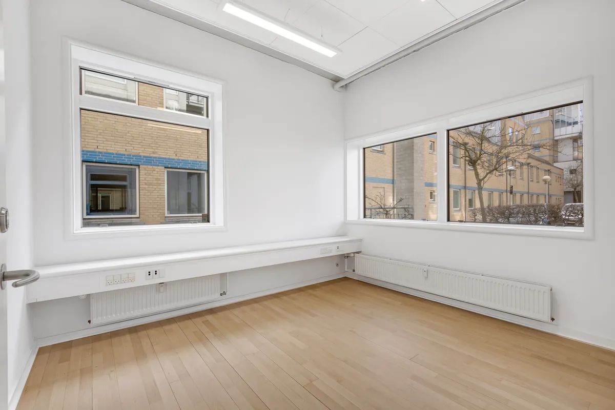 Et moderne kontor med lyse gulve, hvide vægge og store vinduer. Et hvidt radiator er monteret i væggen og en dør er åben.