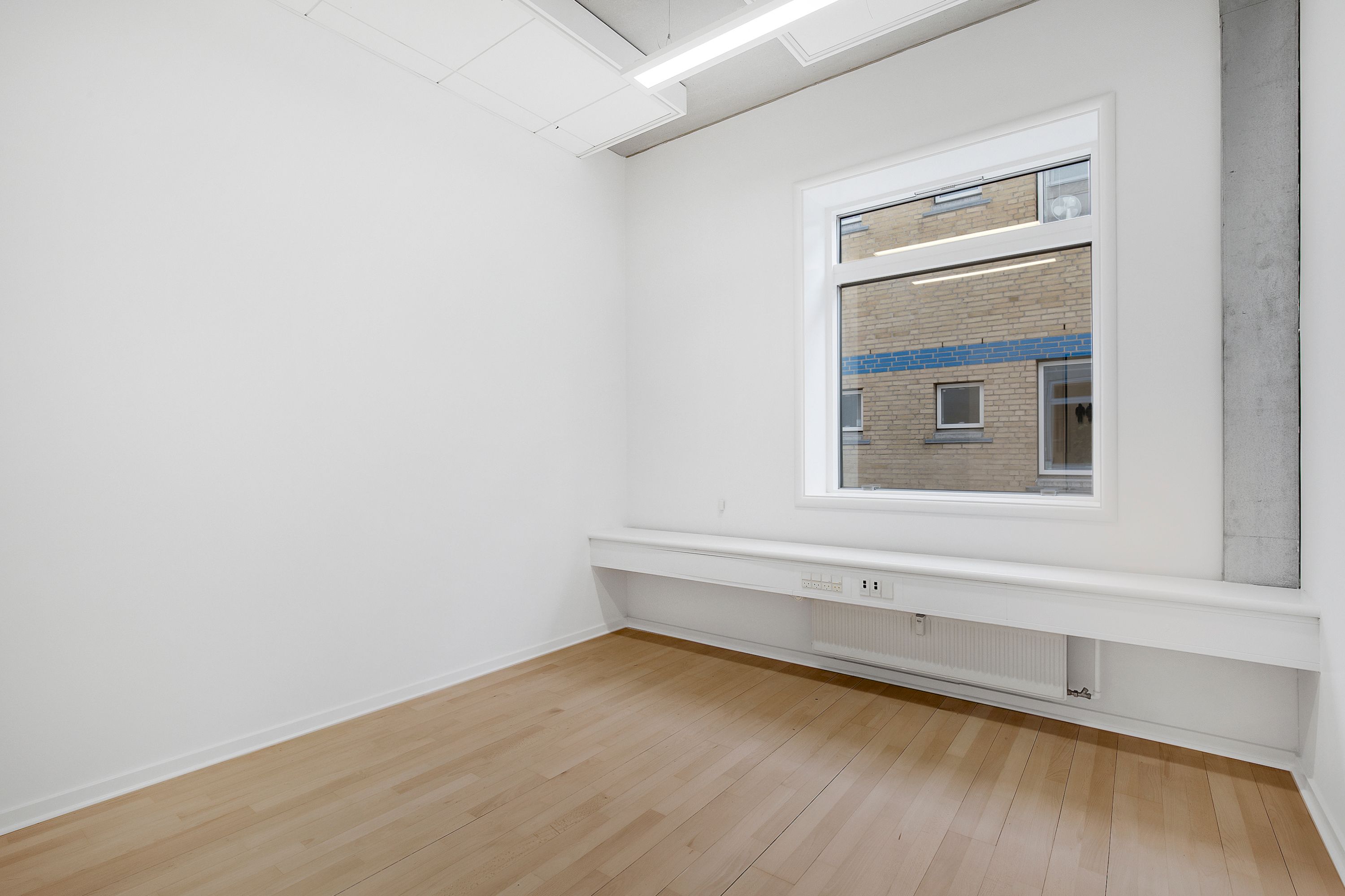 En tom rum med hvide vægge og et vindue med et blik ud til et bygning med røde mursten og blå udsmykning samt et radiator under vinduet
