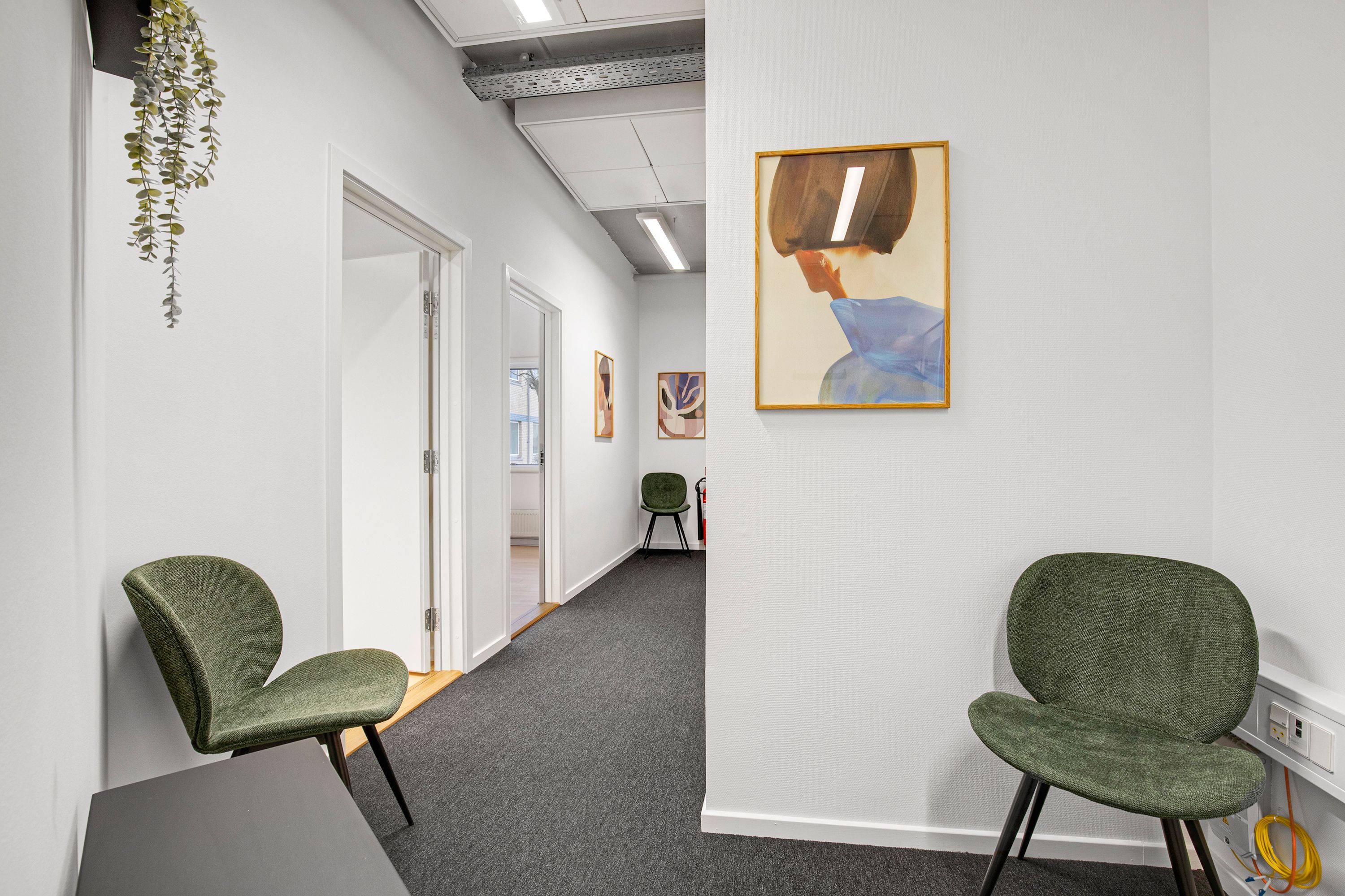 Kontorgang med hvide vægge, plantehænge og kunstværker. To grønne møbler og en slukket brandhøjtalerudgang. Gulvbelægning af gråt tæppe og en skrående væg.