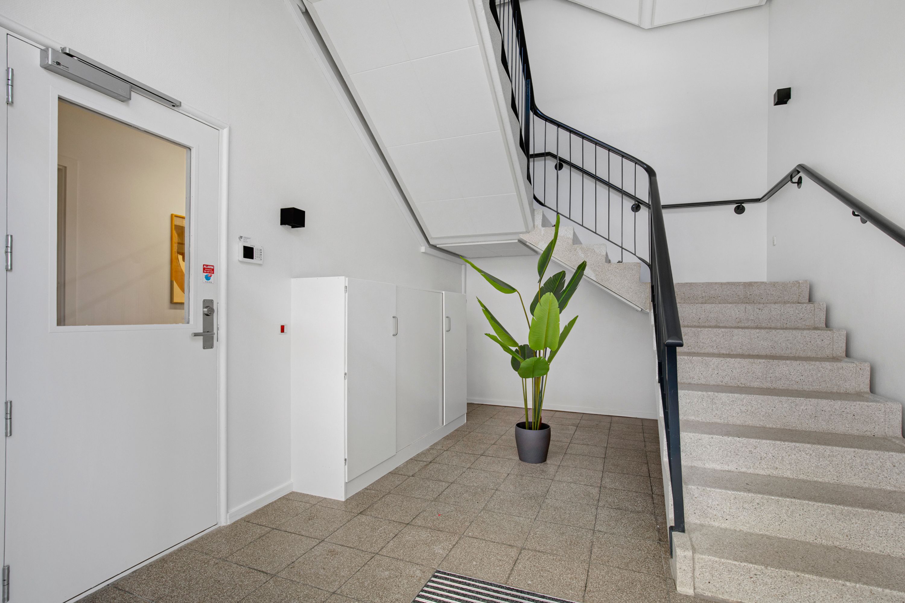 Kontorlokation med hvidt panel og marmortrapper, højt gulv og indgangsdør med automatisk åbner og døre til lagerrum samt plante i gryde