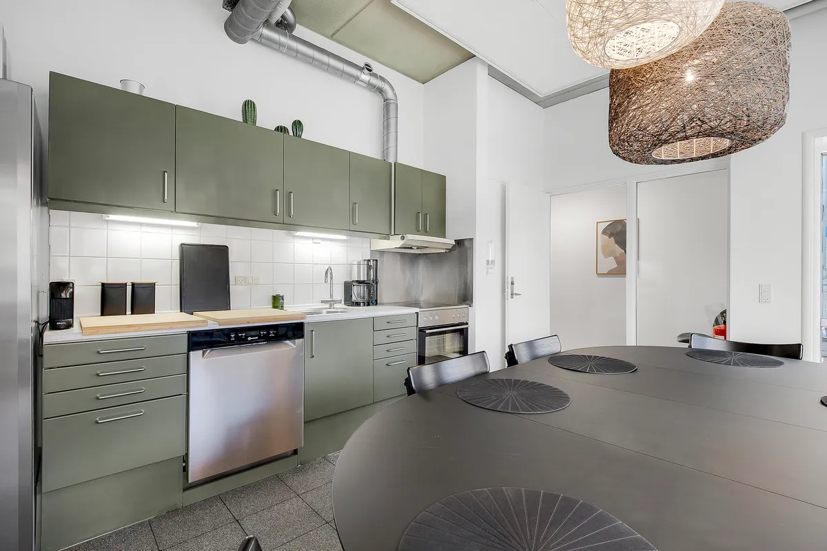 Minimalistisk køkken med en stor rund træbord, moderne apparater og grønne skabe med håndtag, lyse blyvægge og to lyse lysekroner i loftet