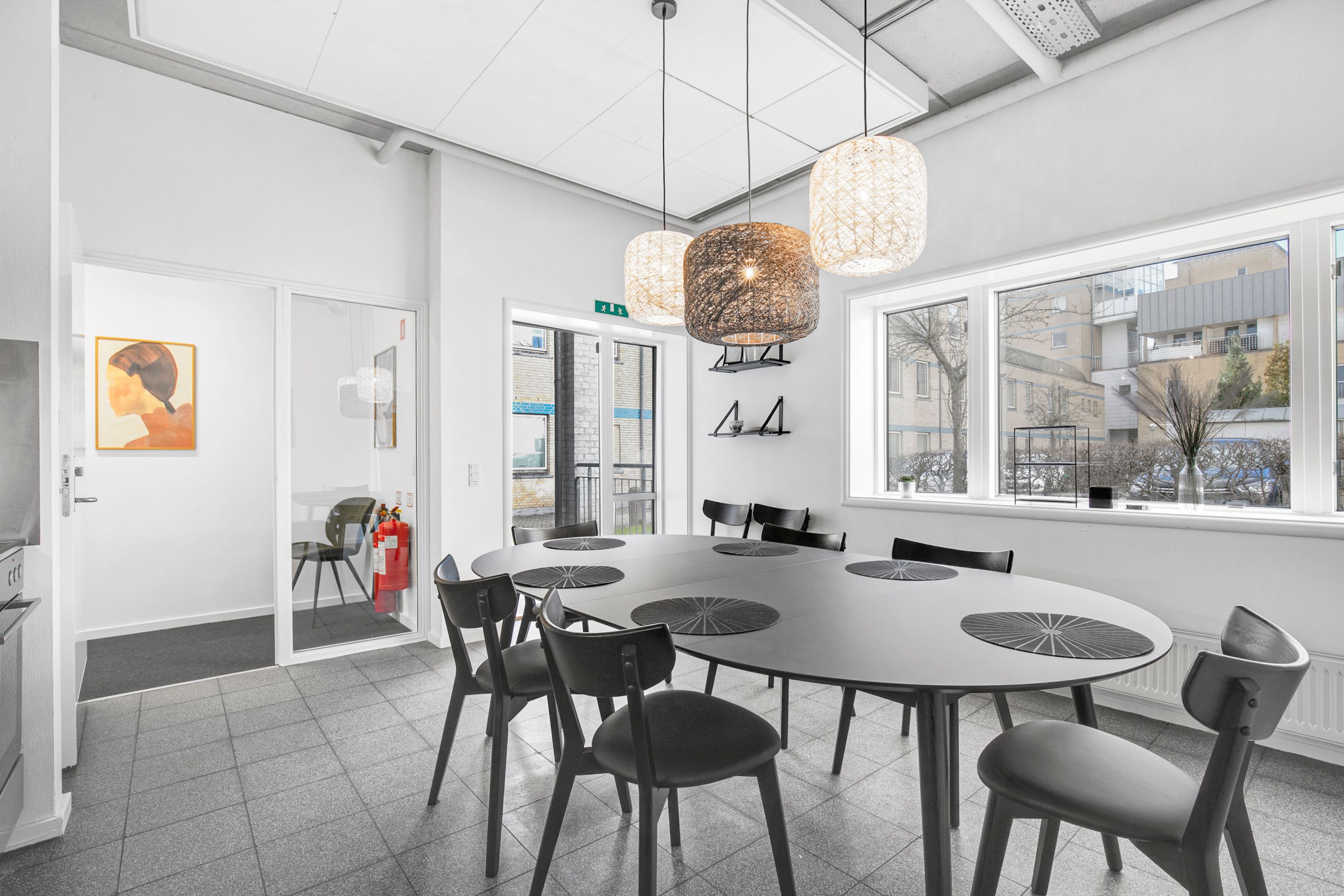 Kontorrum med bord og stole, lysekroner, hvid væg, vinduer, radiator, maleri, og brandværn i nærheden af glasdør