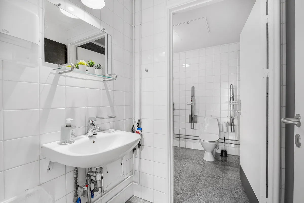 Et moderne badeværelse med hvidt flisebelægning, en hvid vask med håndvask og en torvask, toilet, toiletbørste og en hængende toiletpapirholder